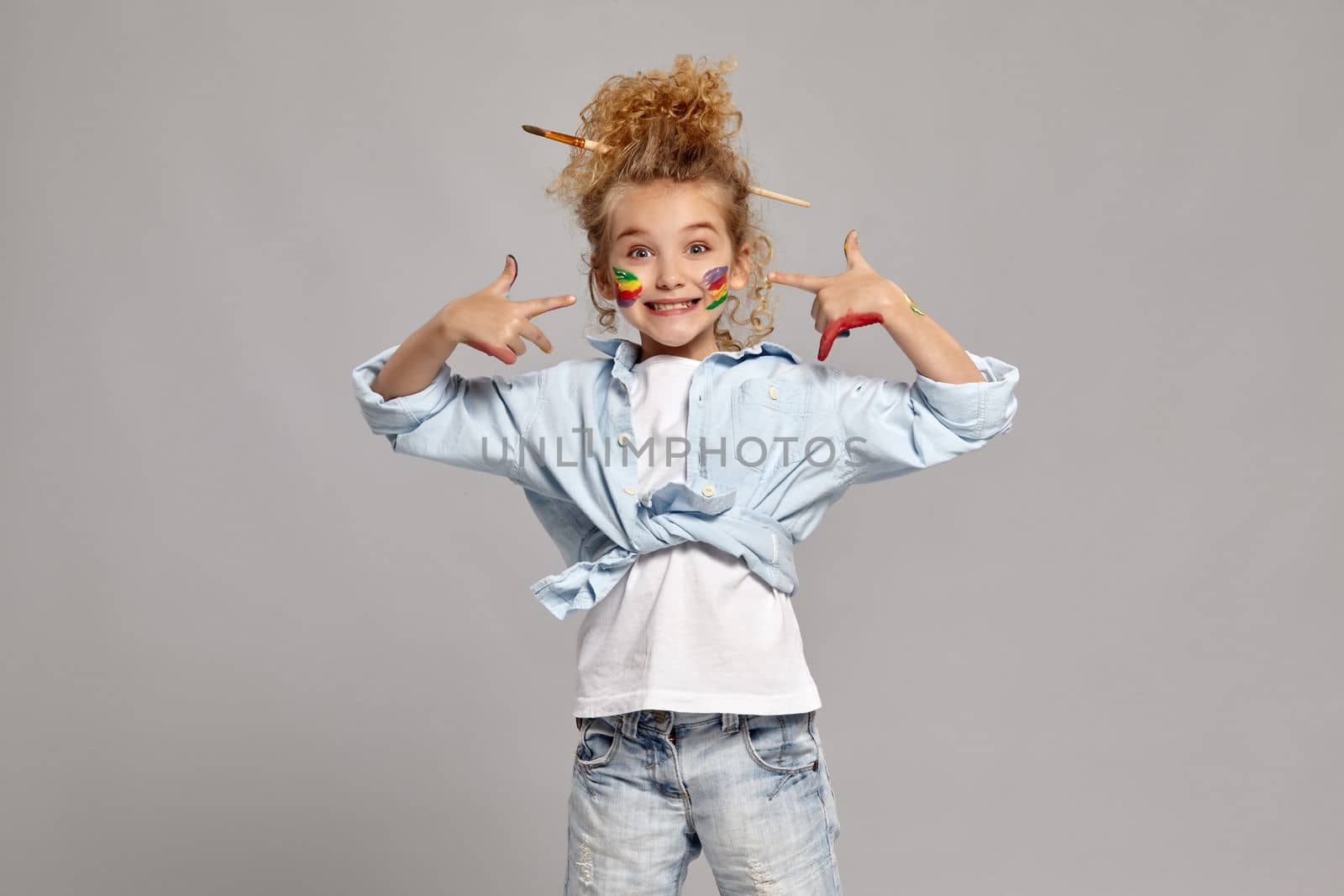 Lovely little lady having a brush in her chic curly blond hair, wearing in a blue shirt and white t-shirt. She has painted her cheeks and pointing on them, smiling at the camera on a gray background.