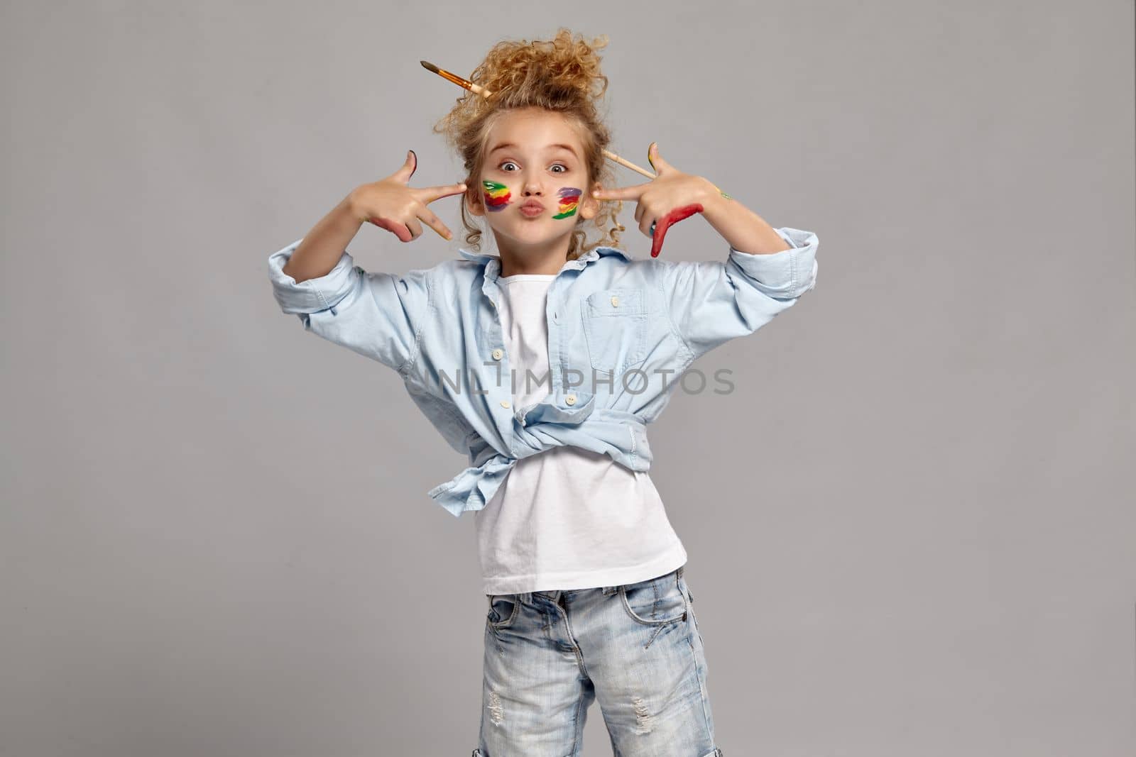 Pretty schoolgirl having a brush in her chic hairstyle, wearing in a blue shirt and white t-shirt. She has painted her cheeks and pointing on them, giving a kiss at the camera on a gray background.