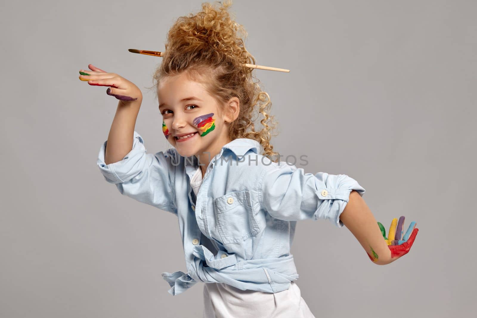 Funny schoolgirl having a brush in her chic curly blond hair, wearing in a blue shirt and white t-shirt. She is posing with a painted hands and cheeks, smiling and looking at the camera, on a gray background.