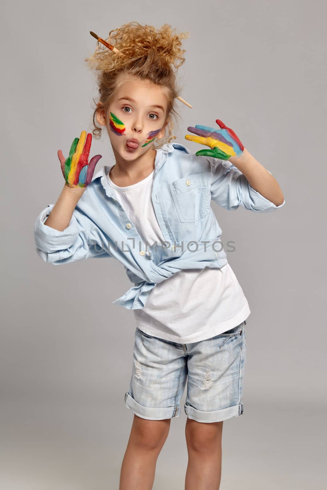 Nice kid having a brush in her chic haircut, wearing in a blue shirt and white t-shirt. She is posing with a painted arms and cheeks, looking at the camera and showing her tongue, on a gray background.