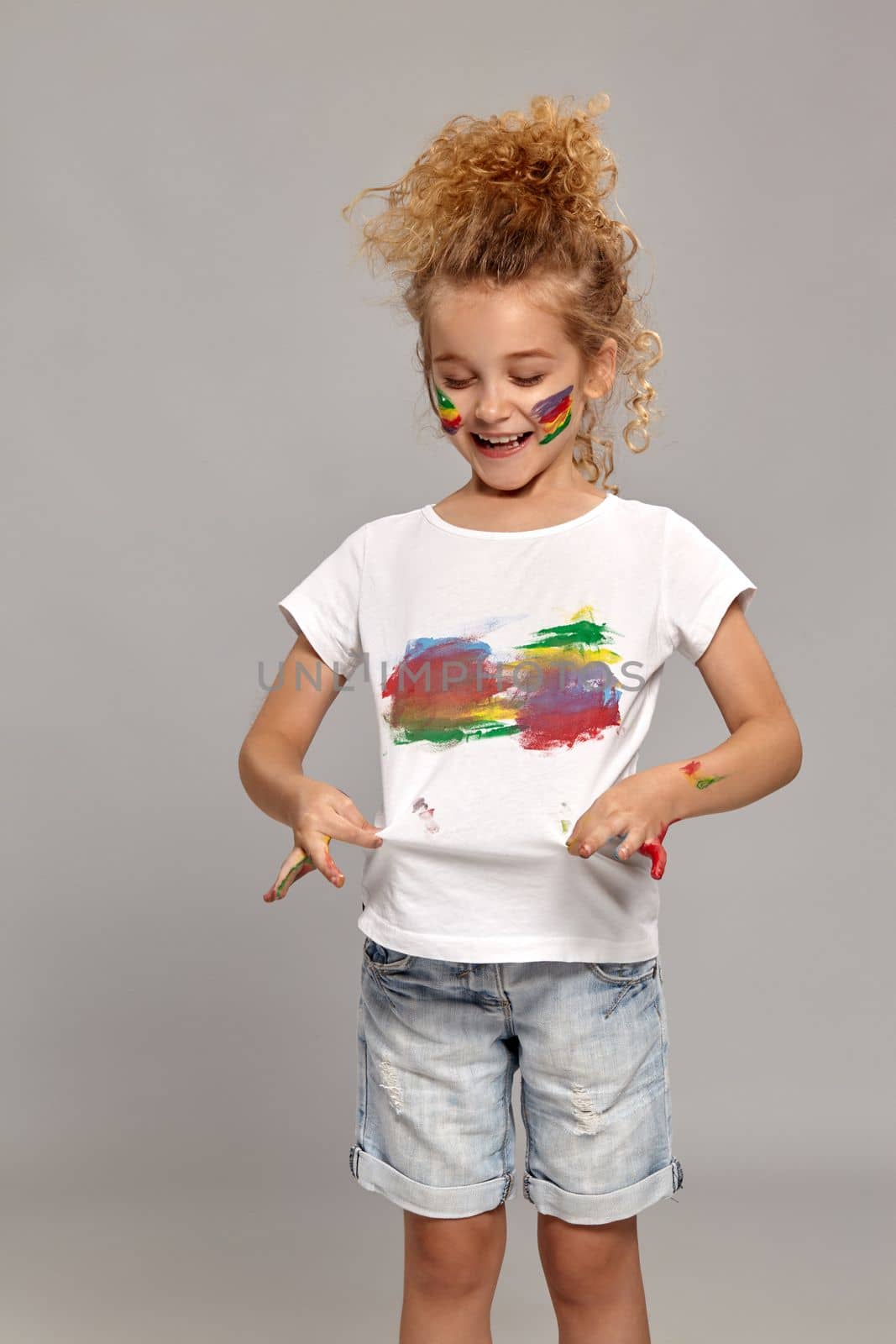 Beautiful little lady having a brush in her lovely haircut, wearing in a white smeared t-shirt. She is looking at it and smiling on a gray background.