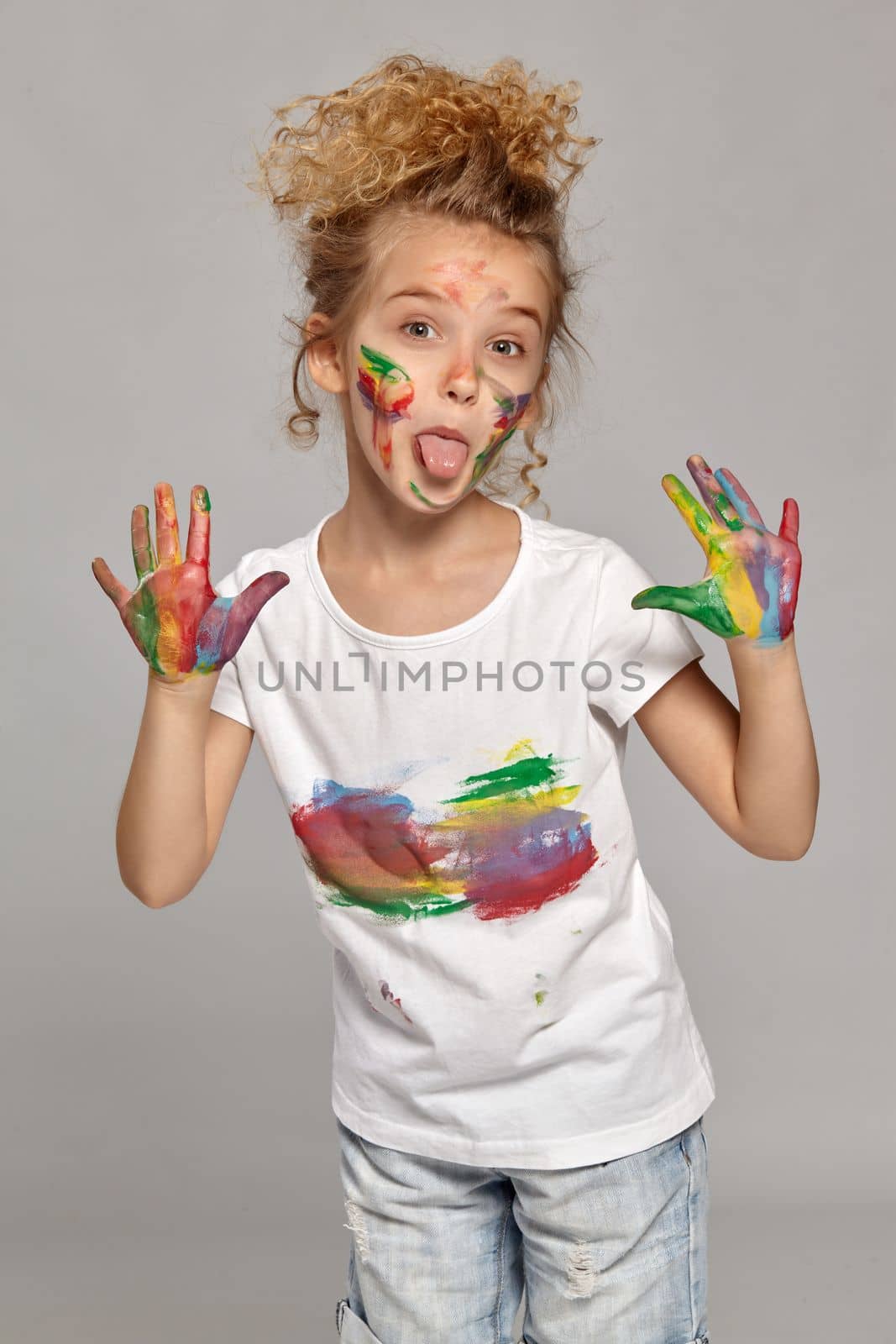 Cute child having a brush in her lovely haircut, wearing in a white smeared t-shirt. She is posing with a painted hands and cheeks and showing her tongue, on a gray background.