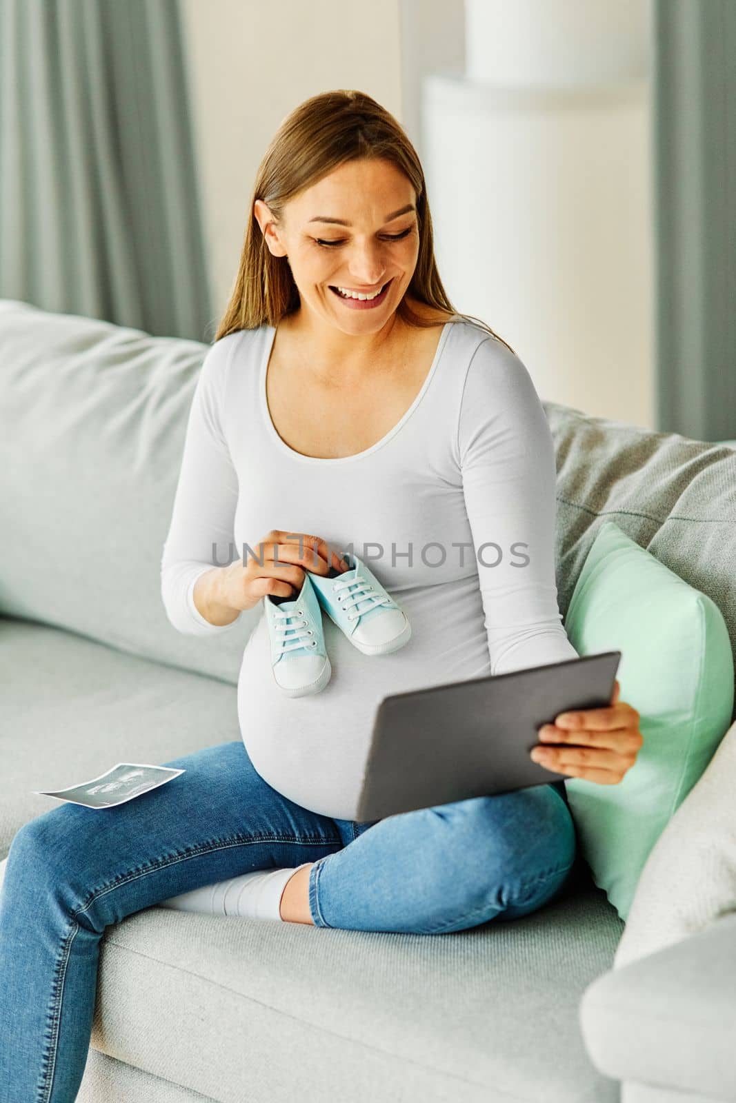 Portrait of a happy young pregnant woman holding small baby shoes and showing on video call on a tablet computer at home