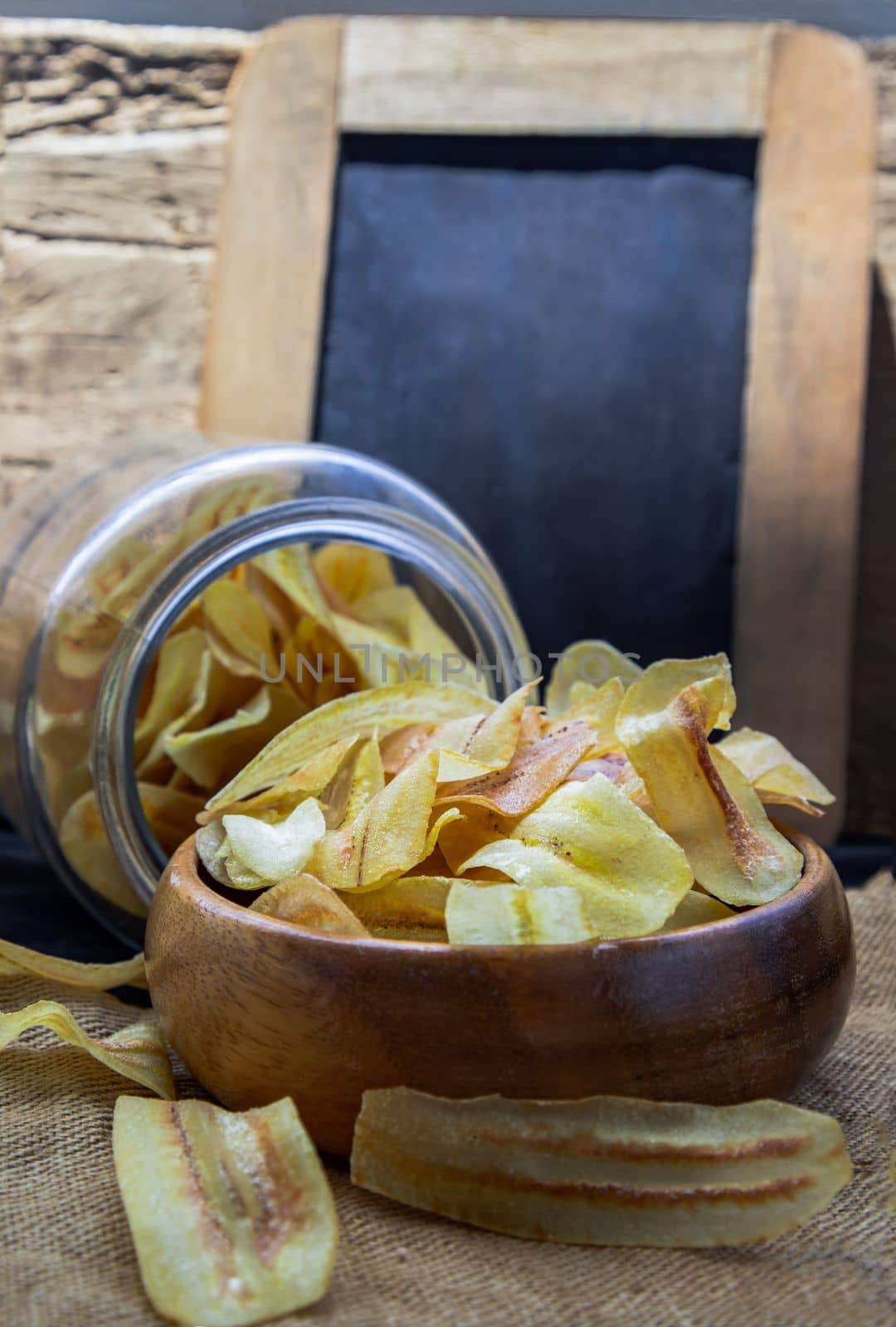 Homemade banana chips in wooden bowl and glass jar on sackcloth with vintage style. Banana dessert, Long banana chips, Empty place for text, Selective focus.