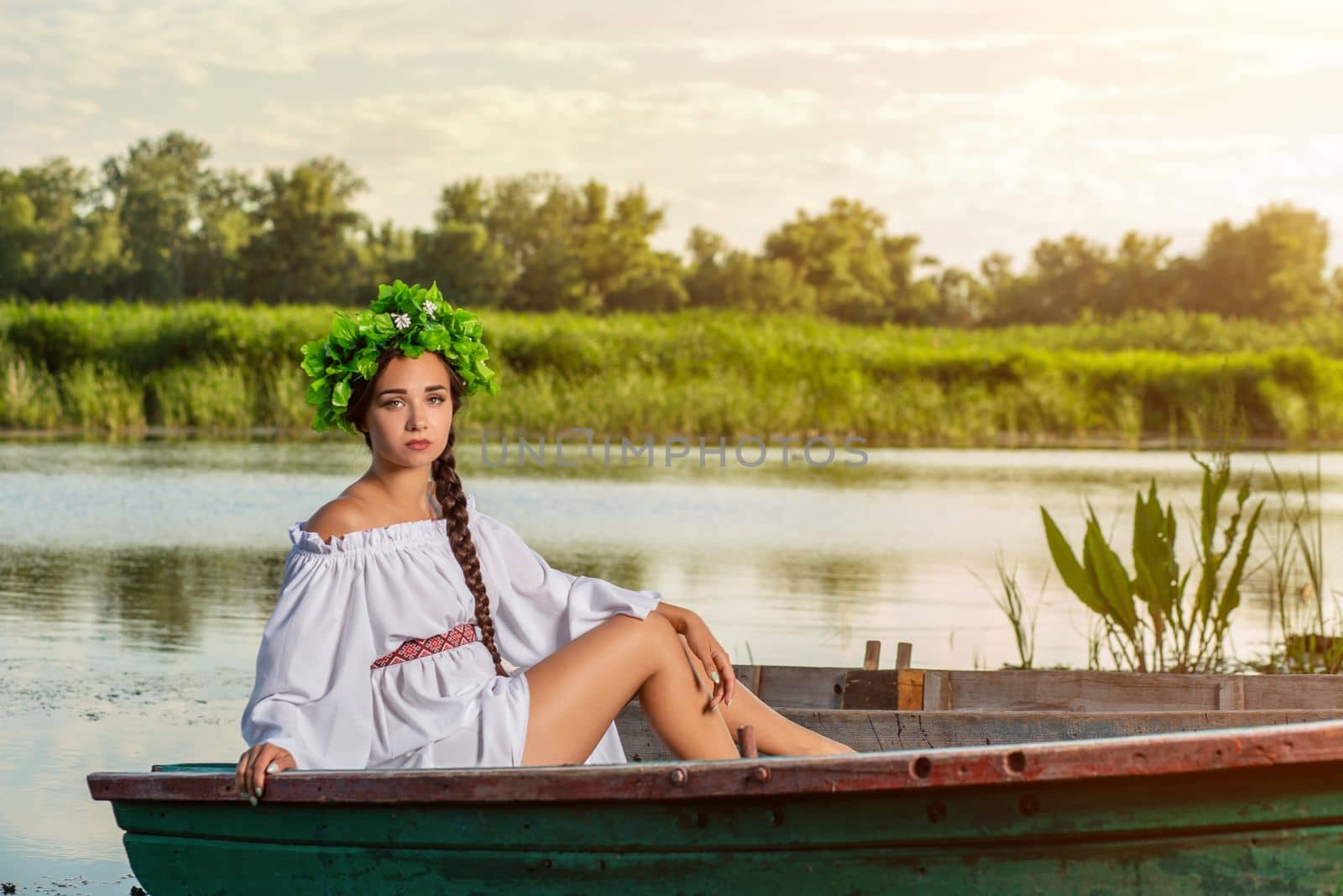 Young sexy woman on boat at sunset. The girl has a flower wreath on her head, relaxing and sailing on river. Beautiful body and face. Fantasy art photography. Concept of female beauty, rest in the village