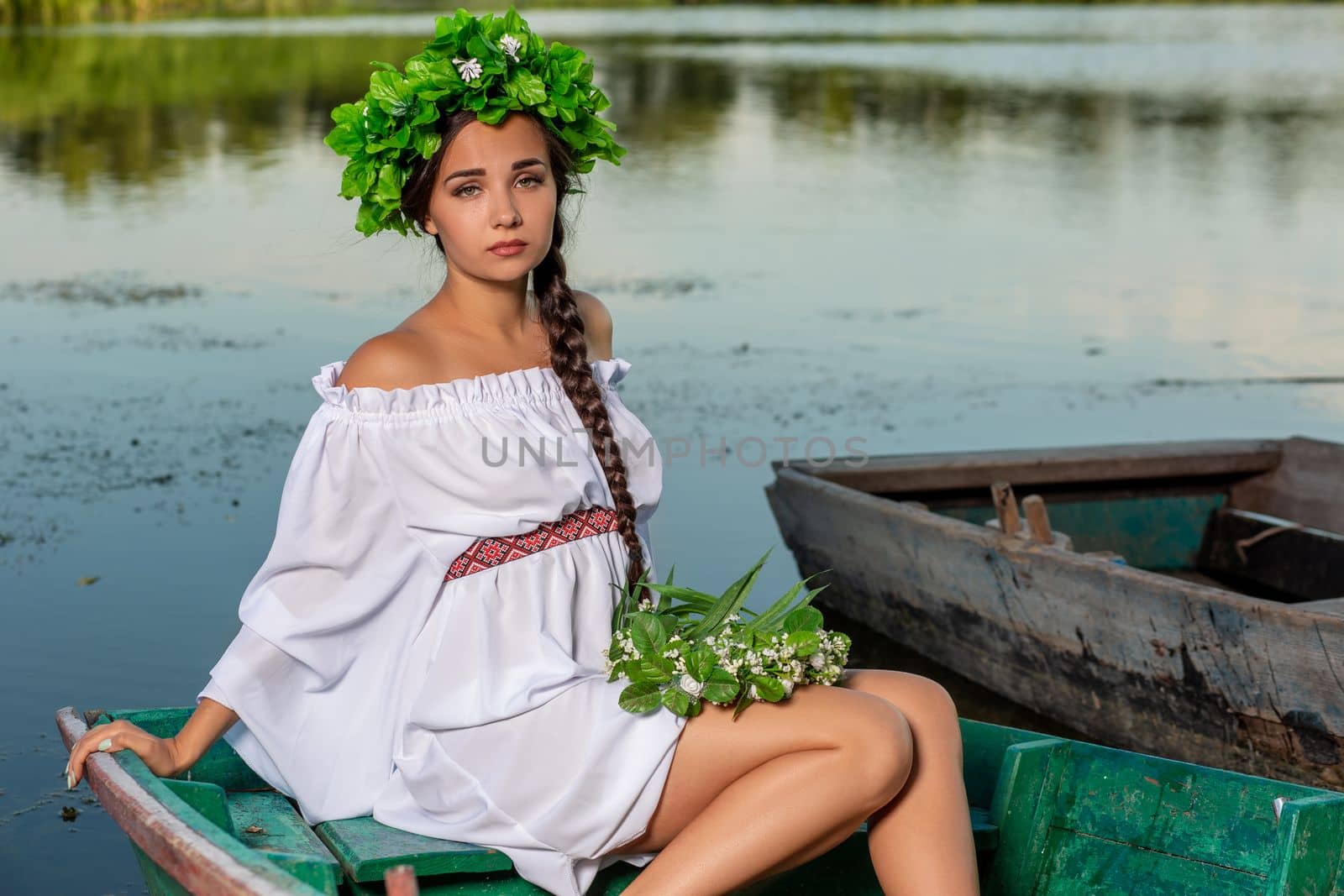 Young sexy woman on boat at sunset. The girl has a flower wreath on her head, relaxing and sailing on river. Beautiful body and face. Fantasy art photography. Concept of female beauty, rest in the village