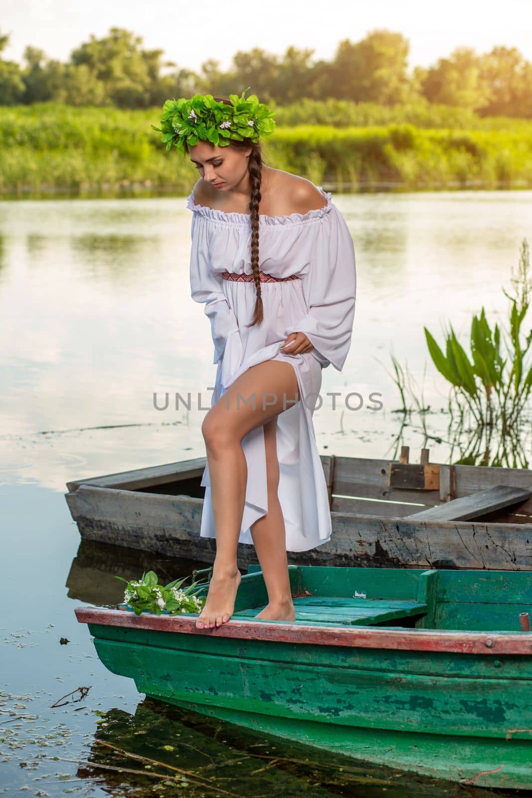 Young sexy woman on boat at sunset. The girl has a flower wreath on her head, relaxing and sailing on river. Beautiful body and face. Fantasy art photography. Concept of female beauty, rest in the village