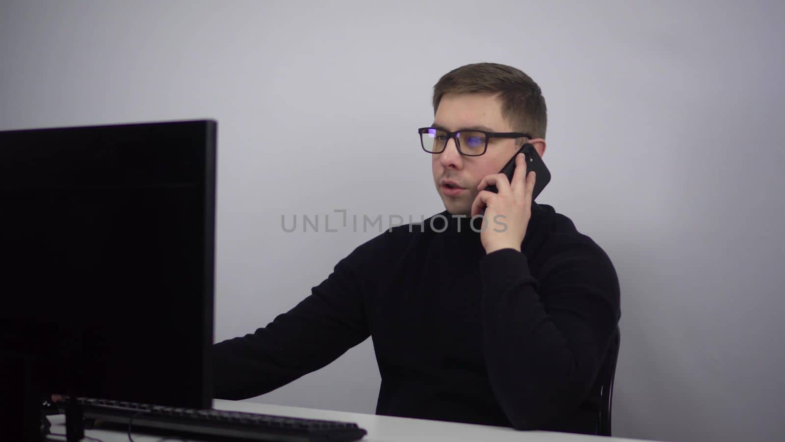 A young man is talking on the phone while sitting at a computer in the office. IT specialist in glasses and a black turtleneck at a remote work. by Puzankov