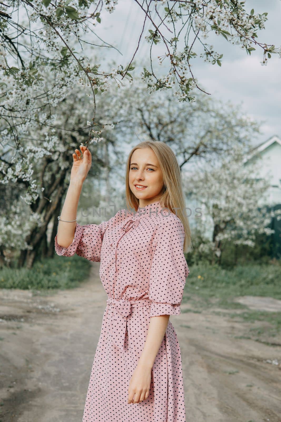 Blonde girl on a spring walk in the garden with cherry blossoms. Female portrait, close-up. A girl in a pink polka dot dress