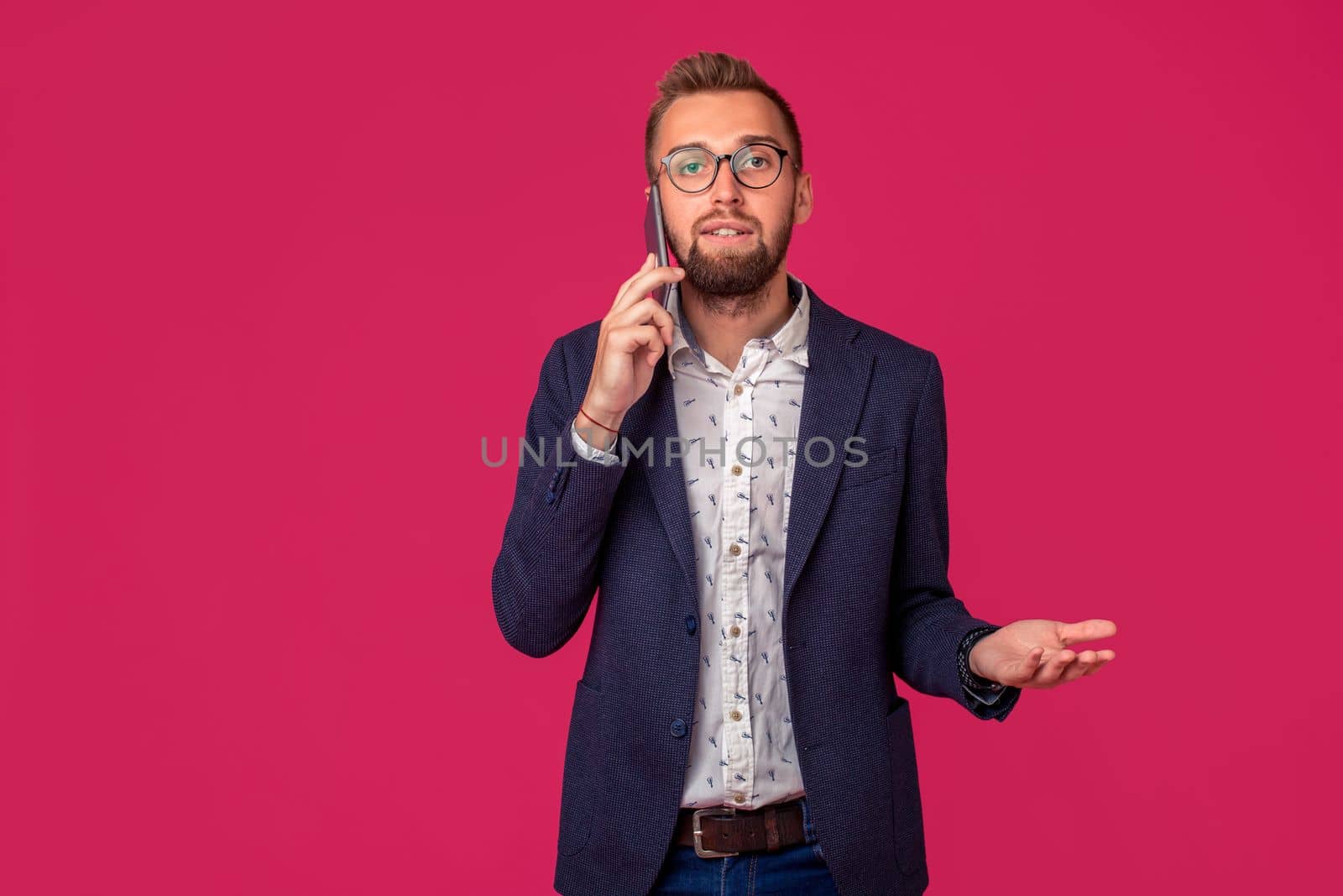 Studio shot of attractive brunette business man with glasses, in casual shirt, stylish black jacket talking on the cell phone. Isolated pink background.