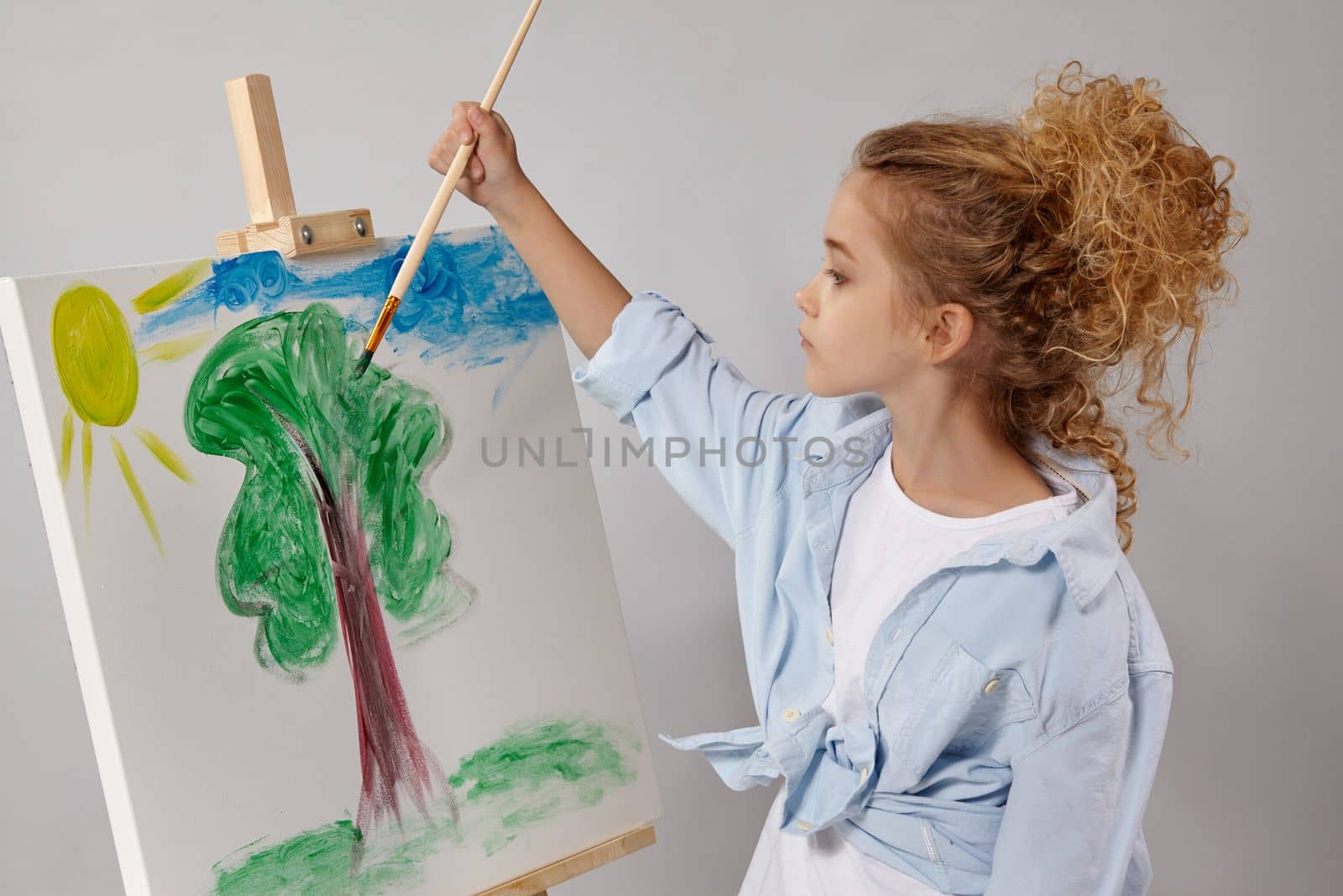 Charming school girl whith a curly blond hair, wearing in a blue shirt and white t-shirt is painting with a watercolor brush on an easel, standing on a gray background.