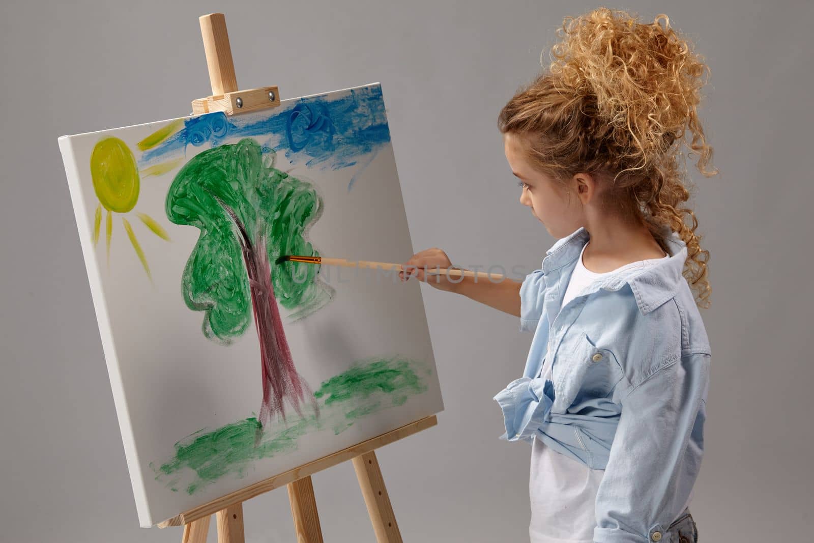 Beautiful school girl whith a curly blond hair, wearing in a blue shirt and white t-shirt is painting with a watercolor brush on an easel, standing on a gray background.