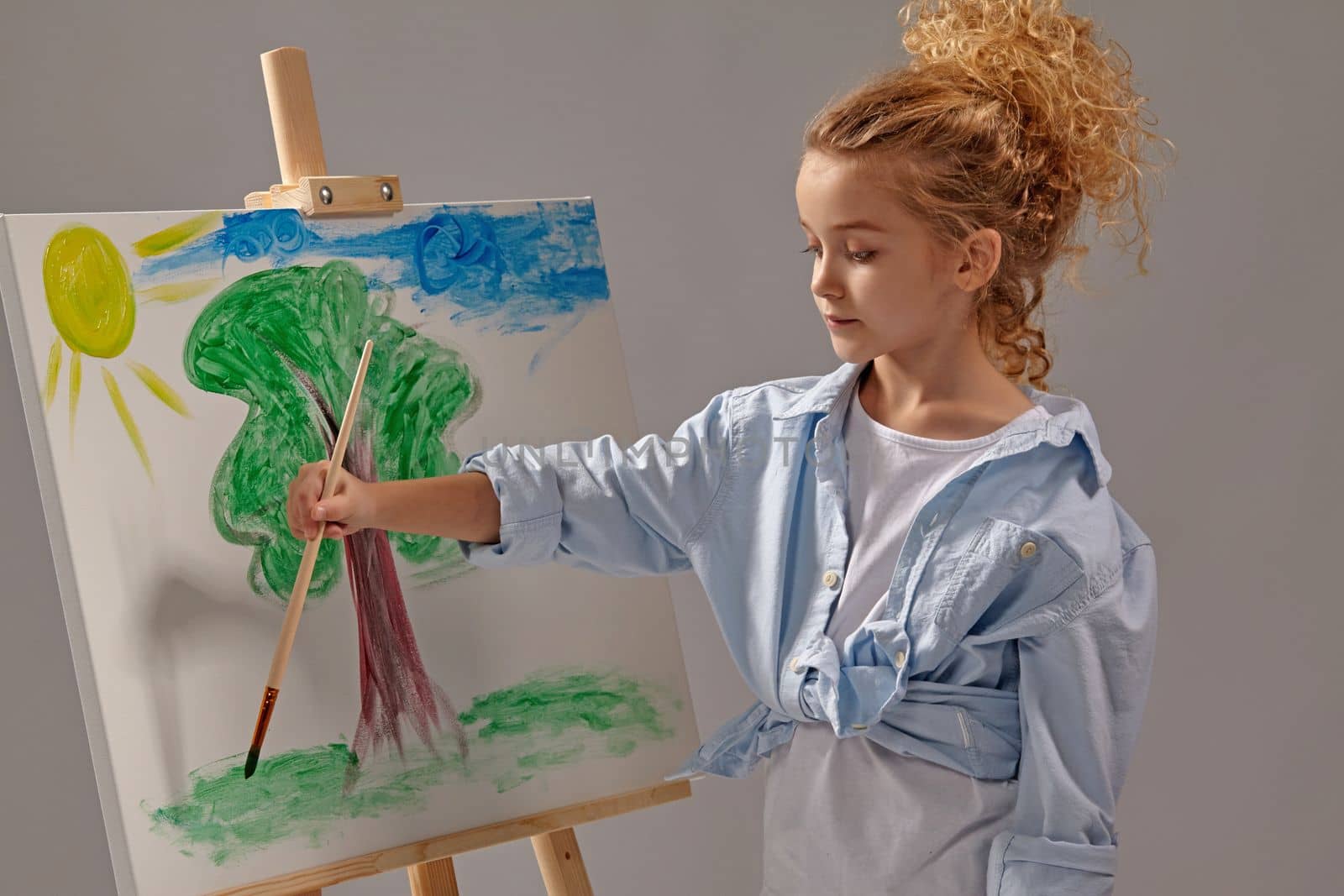 Pretty school girl whith a curly blond hair, wearing in a blue shirt and white t-shirt is painting with a watercolor brush on an easel, standing on a gray background.