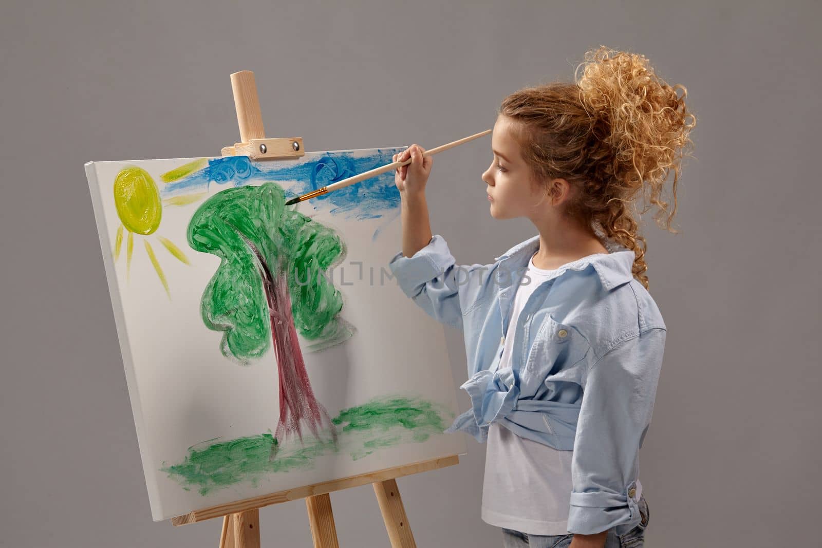 Nice school girl whith a curly blond hair, wearing in a blue shirt and white t-shirt is painting with a watercolor brush on an easel, standing on a gray background.