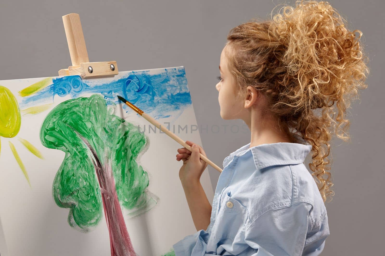 Charming school girl whith a curly hair, wearing in a blue shirt and white t-shirt is painting with a watercolor brush on an easel, standing on a gray background.