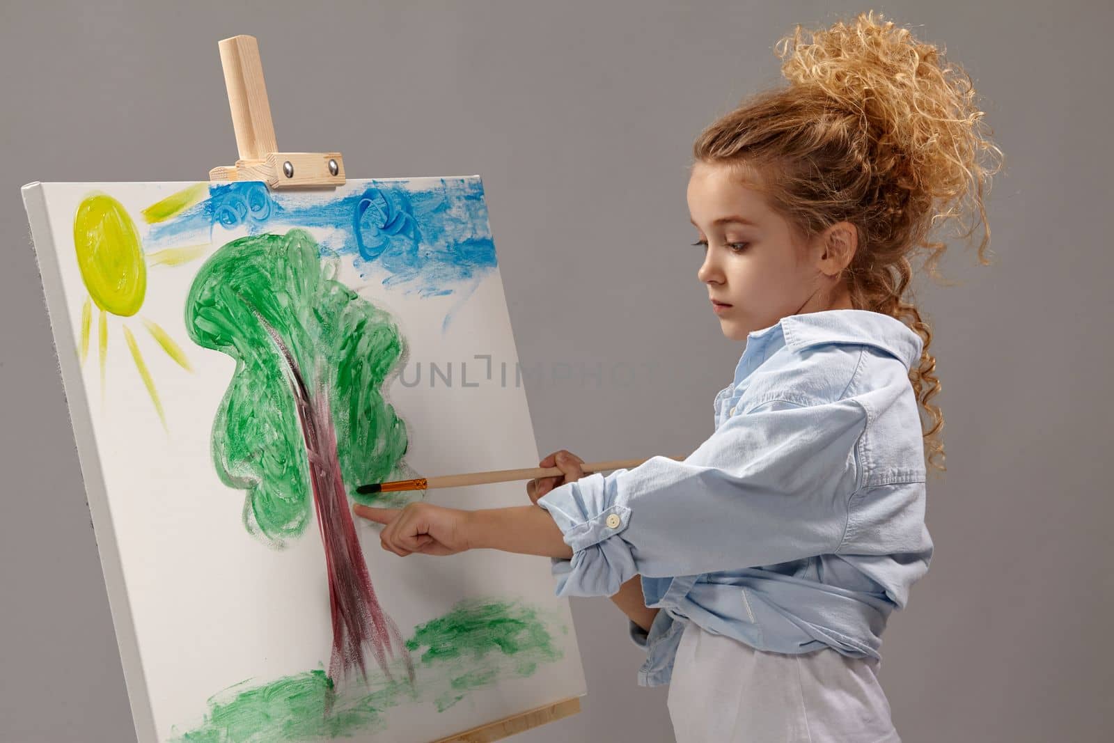 Little school girl whith a curly blond hair, wearing in a blue shirt and white t-shirt is painting with a watercolor brush on an easel, standing on a gray background.