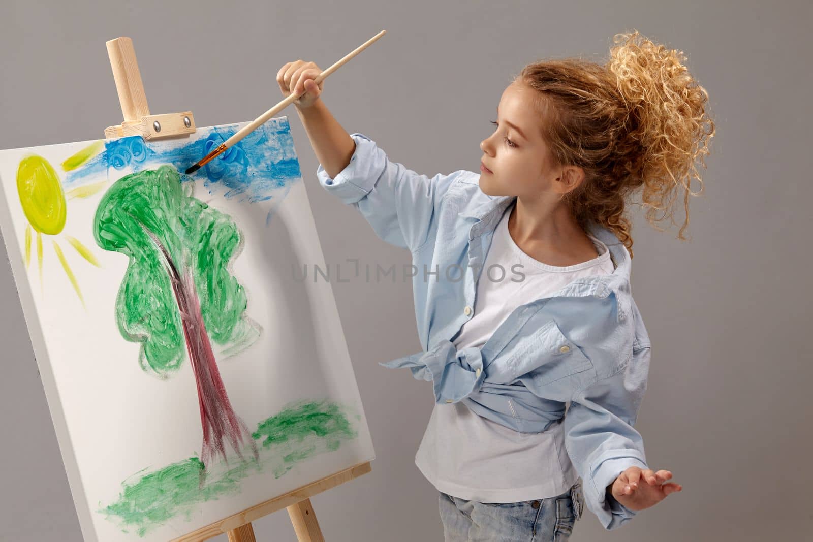 Charming school girl whith a curly hair, wearing in a blue shirt and white t-shirt is painting with a watercolor brush on an easel, standing on a gray background.