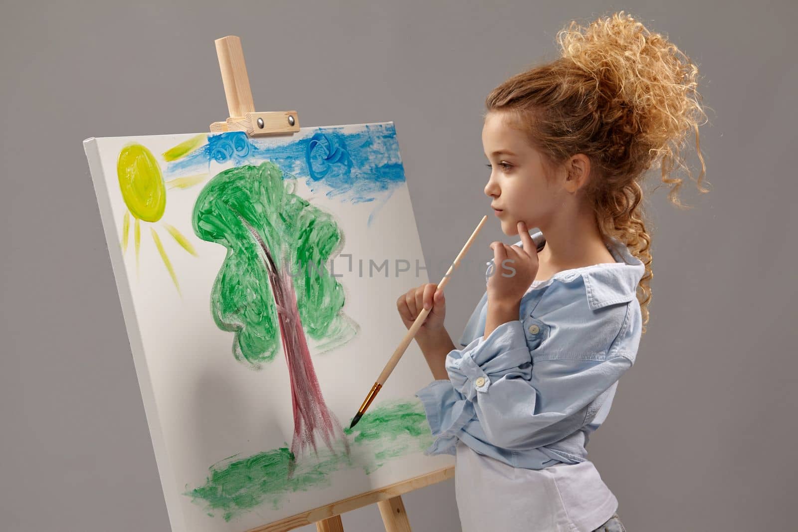 Thoughtful school girl whith a curly blond hair, wearing in a blue shirt and white t-shirt is painting with a watercolor brush on an easel, standing on a gray background.