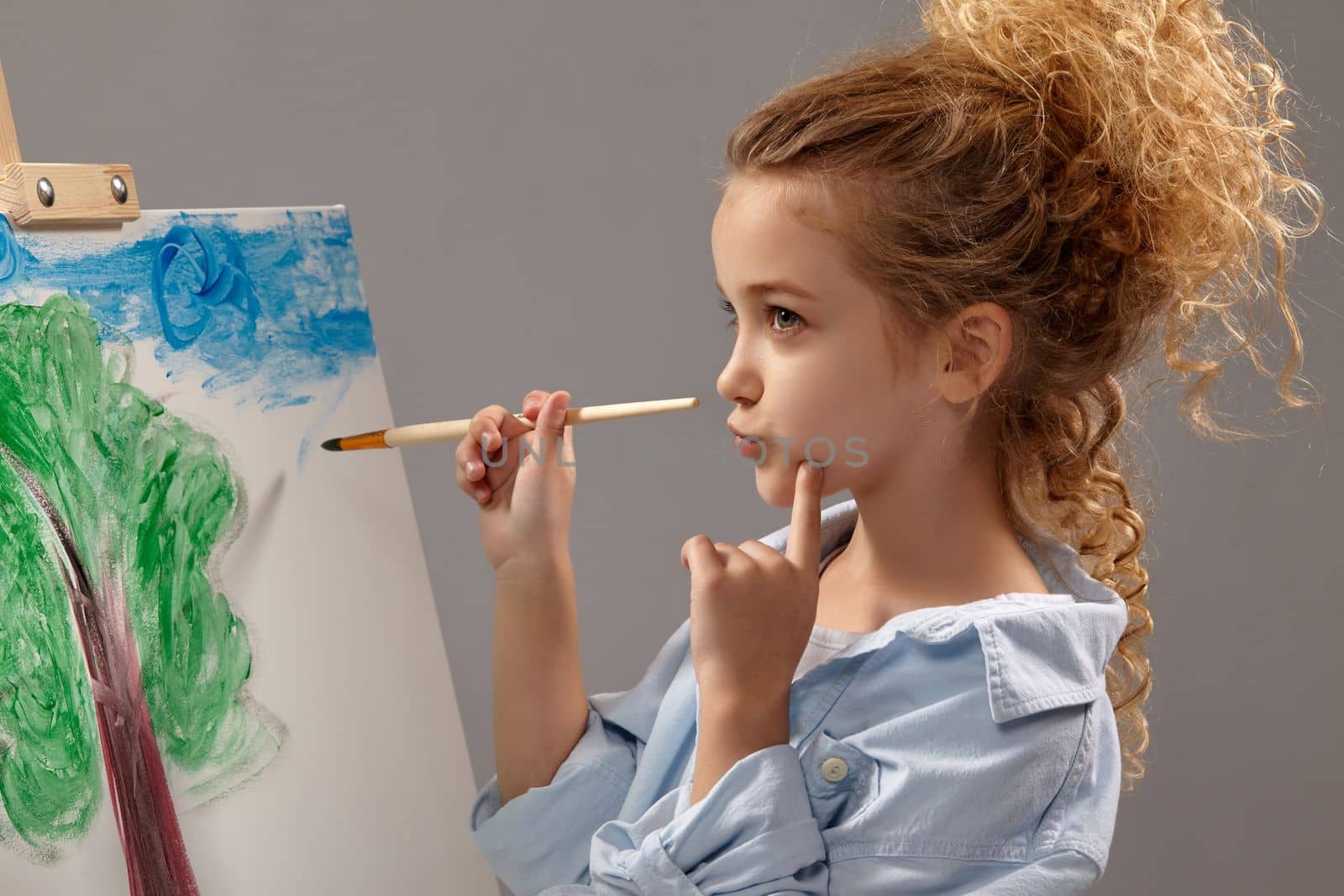 Thoughtful school girl whith a curly hair, wearing in a blue shirt and white t-shirt is painting with a watercolor brush on an easel, standing on a gray background.