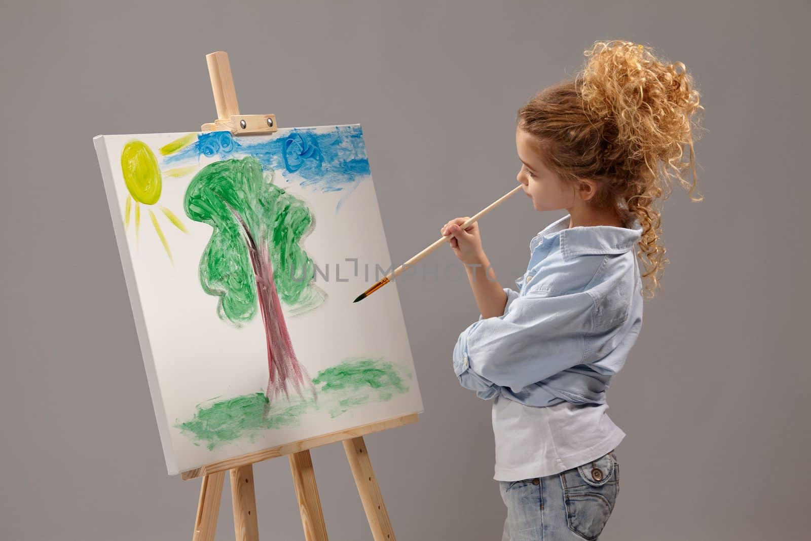 Talanted school girl whith a curly blond hair, wearing in a blue shirt and white t-shirt is painting with a watercolor brush on an easel, standing on a gray background.