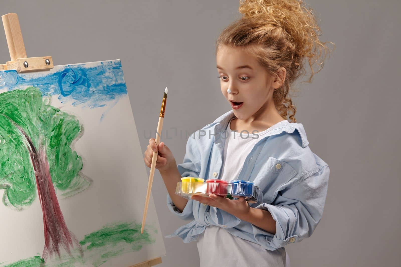Talanted school girl whith a curly blond hair, wearing in a blue shirt and white t-shirt is painting with a watercolor brush on an easel, looking wondered and standing on a gray background.