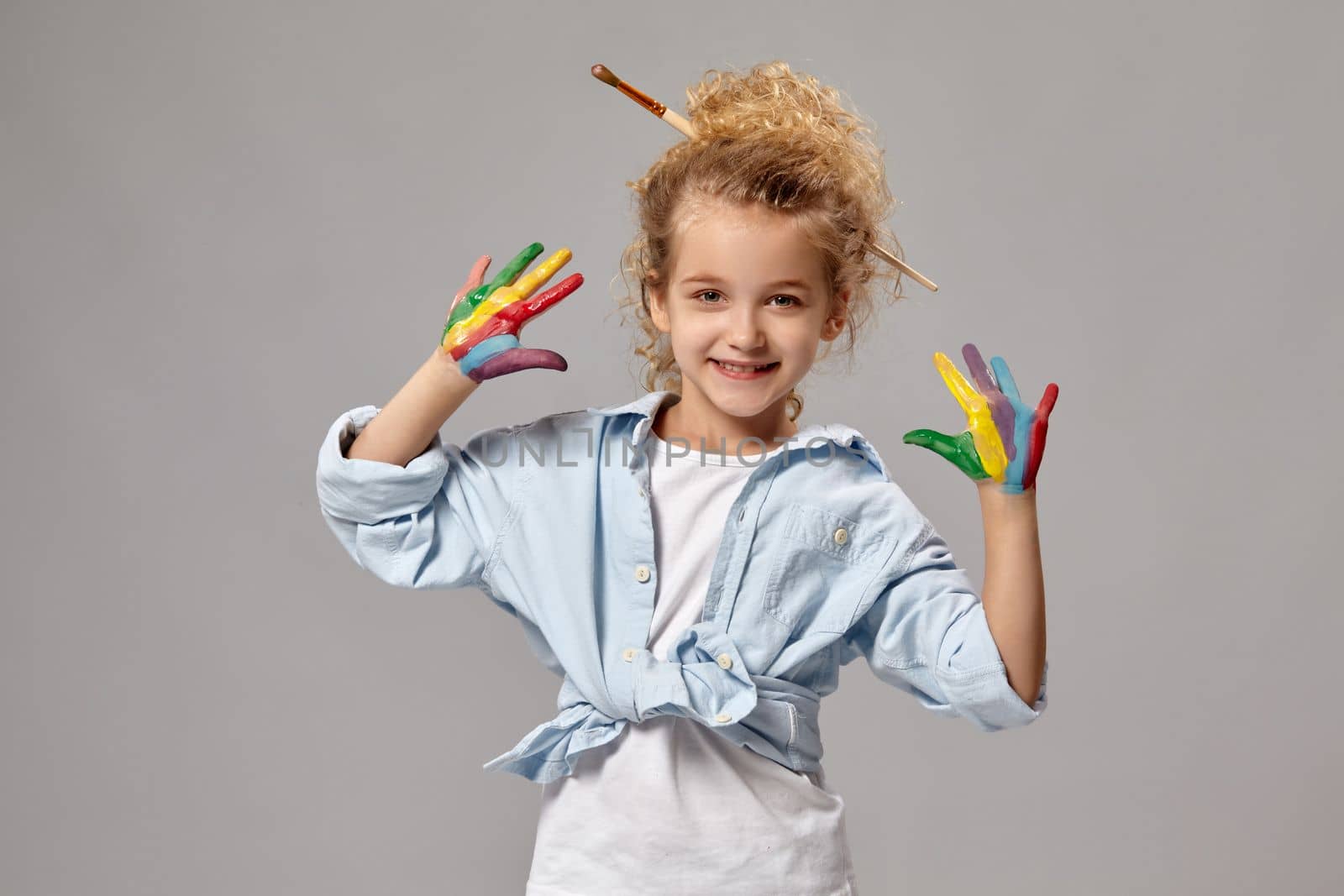 Beautiful little girl having a brush in her chic curly blond hair, wearing in a blue shirt and white t-shirt, with a painted fingers is posing on a gray background.