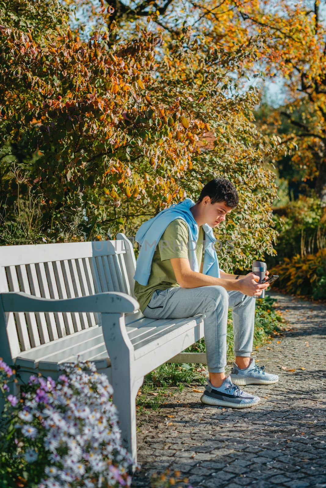 A Teenager Sits On A Bench In The Autumn Park Drinks Coffee From A Thermo Mug And Looks Into A Phone. Portrait Of Handsome Cheerful Guy Sitting On Bench Fresh Air Using Device Browsing Media Smm Drinking Latte Urban Outside Outdoor by Andrii_Ko