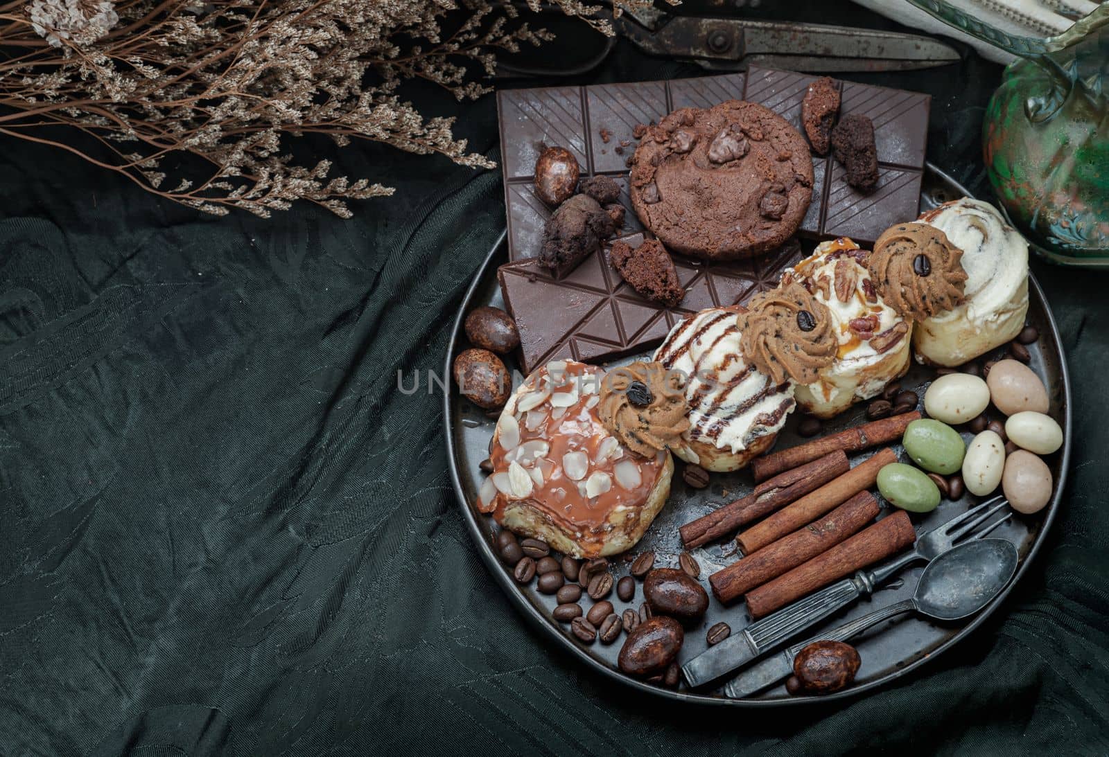 Assorted Cinnamon Rolls, Dark chocolate bars, cookies, pieces and balls served with black iron cutlery in black ceramic plate on dark background. Top view, Space for text, Selective