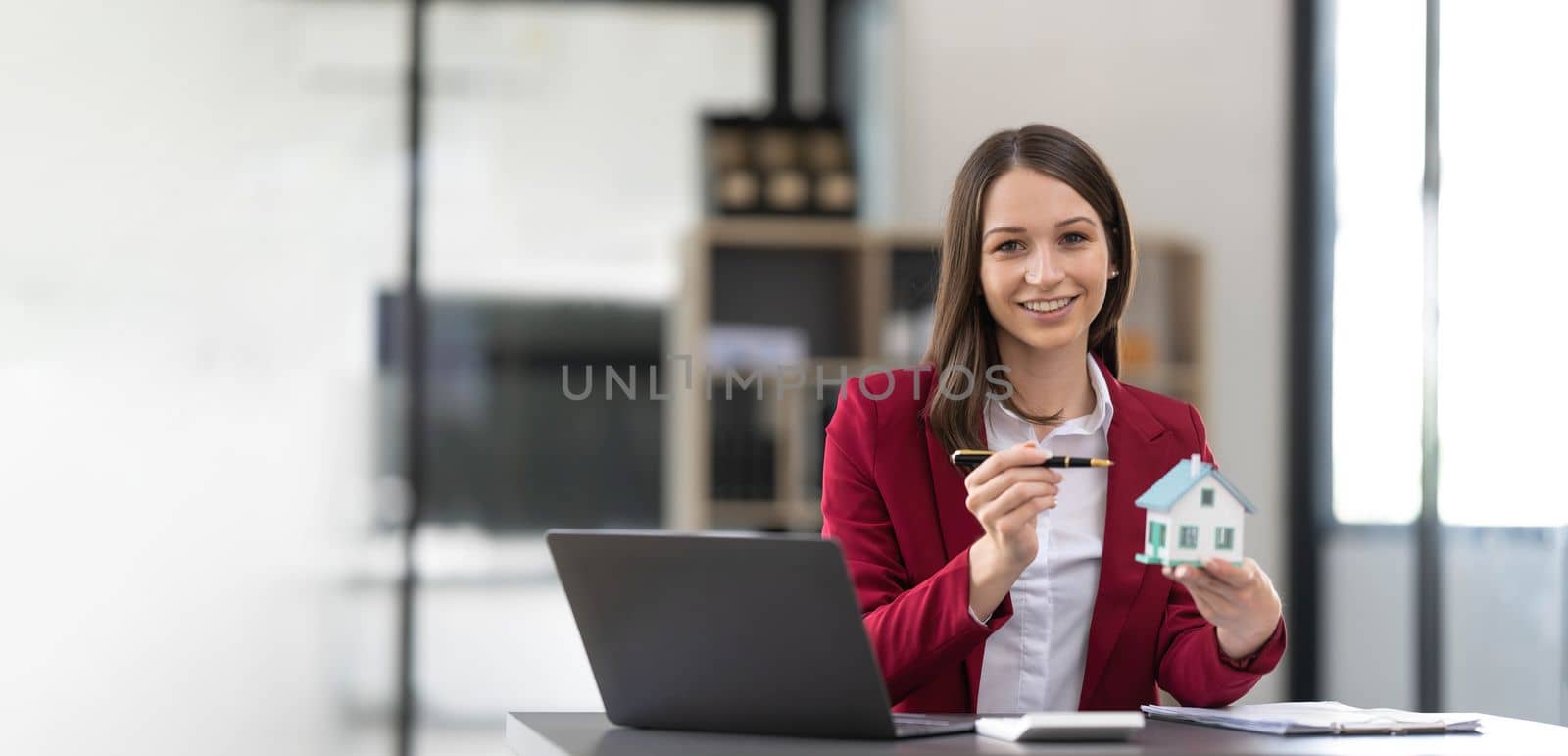 Young Asian real estate agent worker working with laptop at table in office and small house beside it. by wichayada