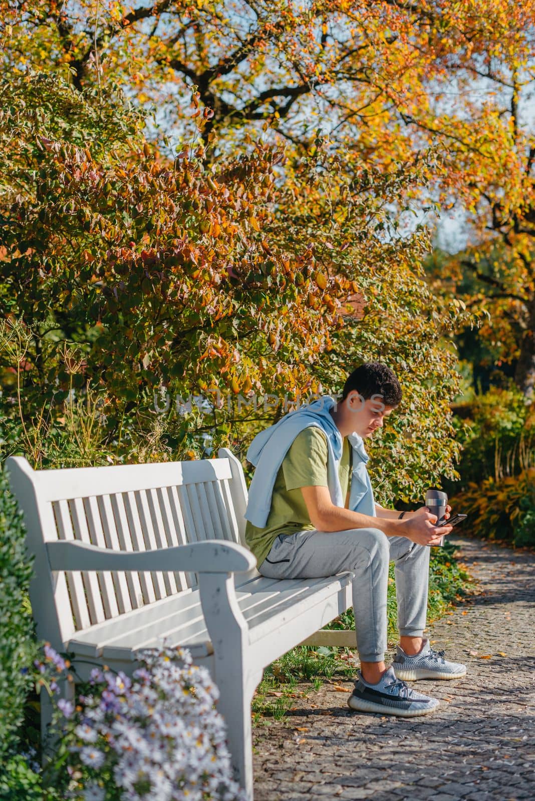 A Teenager Sits On A Bench In The Autumn Park Drinks Coffee From A Thermo Mug And Looks Into A Phone. Portrait Of Handsome Cheerful Guy Sitting On Bench Fresh Air Using Device Browsing Media Smm Drinking Latte Urban Outside Outdoor by Andrii_Ko