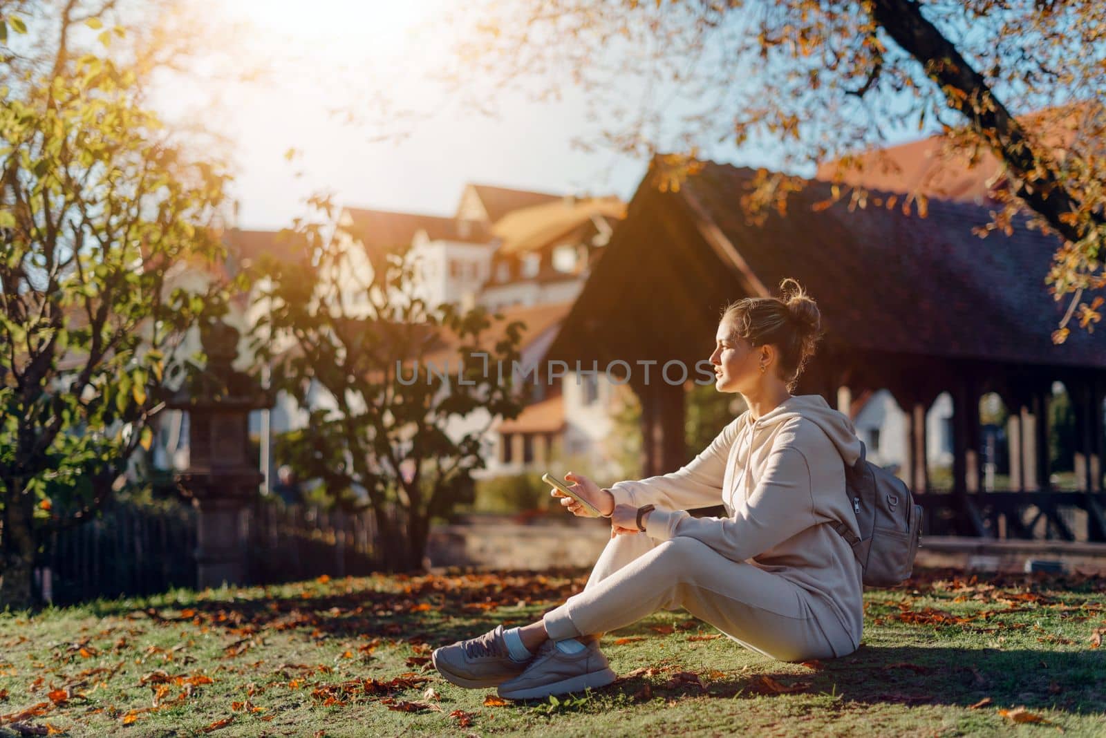 Young fashionable teenage girl with smartphone in park in autumn sitting at smiling. Trendy young woman in fall in park texting. Retouched, vibrant colors. Beautiful blonde teenage girl wearing casual modern autumn outfit sitting in park in autumn. Retouched, vibrant colors, brownish tones.