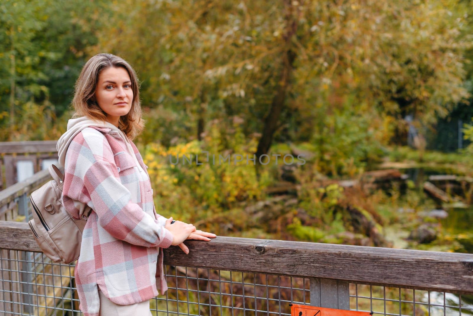 Portrait Of Cute Young Woman In Casual Wear In Autumn, Standing On Bridge Against Background Of An Autumn Park And River. Pretty Female Walking In Park In Golden Fall. Copy Space. Smiling Girl In The Park Standing On Wooden Bridge And Looking At The Camera In Autumn Season by Andrii_Ko