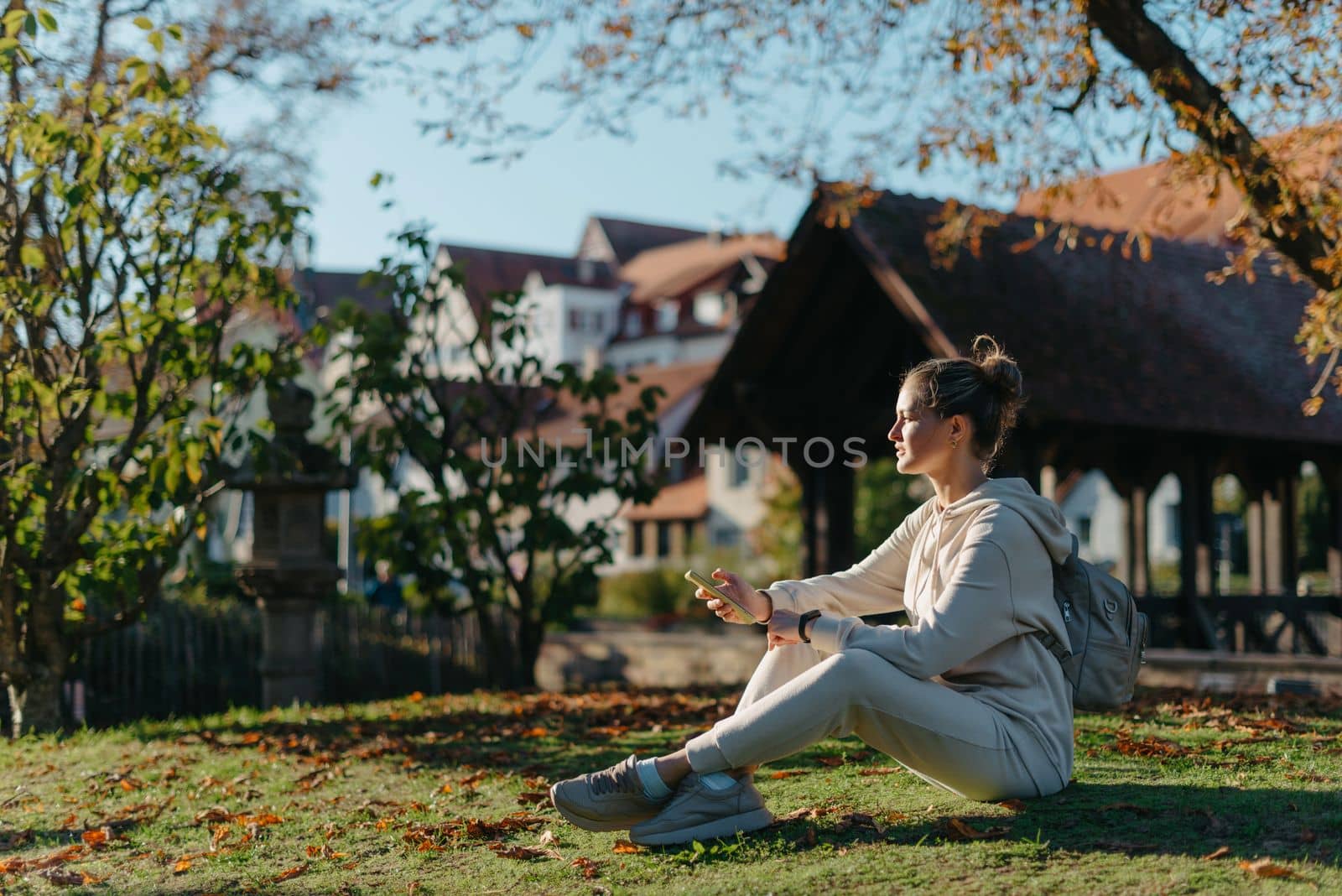 Young Fashionable Teenage Girl With Smartphone In Europian Park In Autumn Sitting At Smiling. Trendy Young Woman In Fall In Park Texting. Retouched, Vibrant Colors. Beautiful Blonde Teenage Girl Wearing Casual Modern Autumn Outfit Sitting In Park In Autumn. Retouched, Vibrant Colors, Brownish Tones. by Andrii_Ko