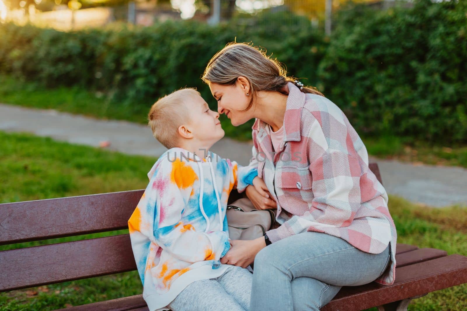 mother and son sit on a park bench in the rays of the setting sun. the concept of a family. Mother's Day. beautiful girl (mother) with a boy (son) in the park in the park are sitting on a bench at sunset by Andrii_Ko