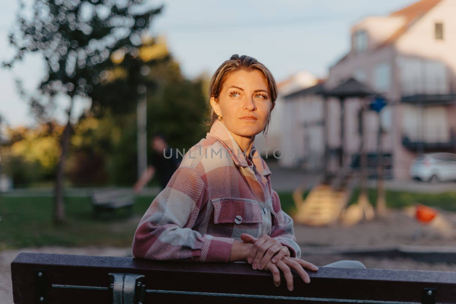 Girl enjoying city view from a bench in sunset or sunrise time. Beautiful blond-haired pretty girl in summer green park against sunset beams. Lonely woman sitting alone moments sunset. On Blurred background bokeh. light Fair. Girl sitting on a bench and watching distant city scenic. by Andrii_Ko