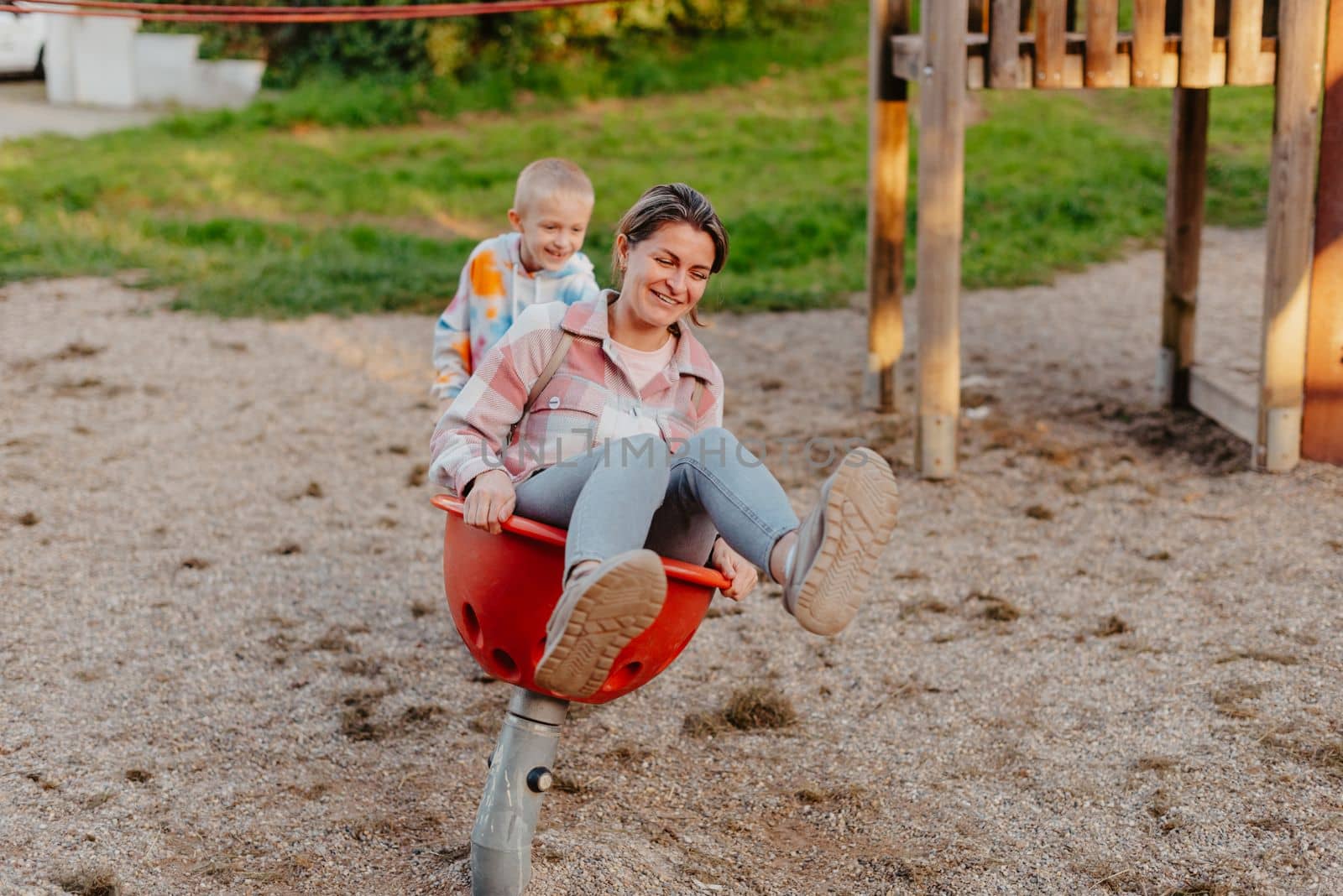 Mother and son having fun on a swing. Motherhood and childhood concept. Sunny summer day. Beautiful family is having fun outside. Parents with children riding on a swing. Mom is playing with her little son on a terrace with swings. A mother woman swings on a swing in the park. Mom and child are on a swing in nature by Andrii_Ko