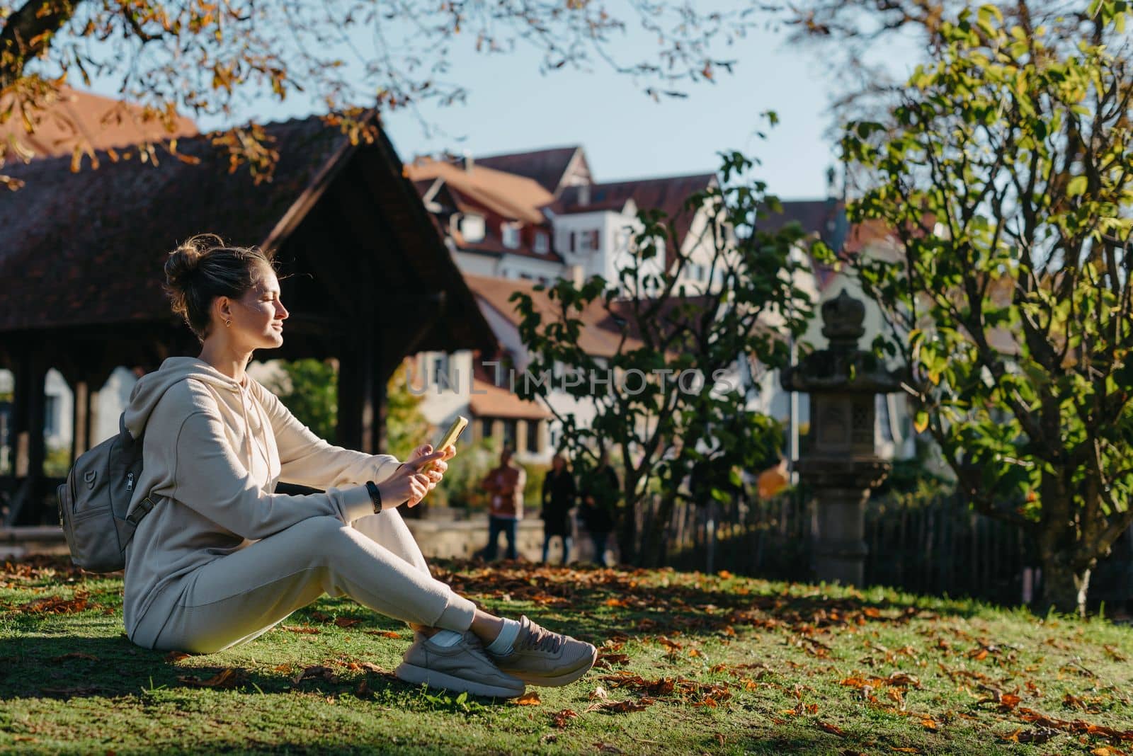 Young fashionable teenage girl with smartphone in park in autumn sitting at smiling. Trendy young woman in fall in park texting. Retouched, vibrant colors. Beautiful blonde teenage girl wearing casual modern autumn outfit sitting in park in autumn. Retouched, vibrant colors, brownish tones.