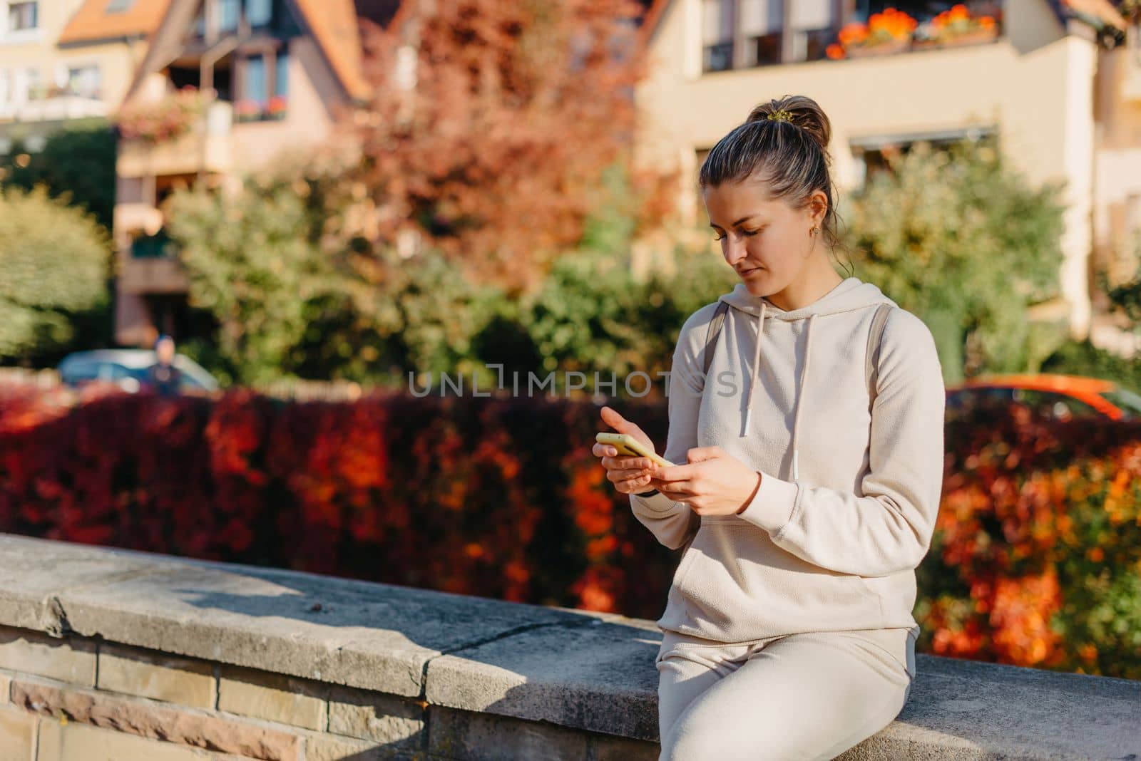 Young Fashionable Teenage Girl With Smartphone In Europian Park In Autumn Sitting At Smiling. Trendy Young Woman In Fall In Park Texting. Retouched, Vibrant Colors. Beautiful Blonde Teenage Girl Wearing Casual Modern Autumn Outfit Sitting In Park In Autumn. Retouched, Vibrant Colors, Brownish Tones. by Andrii_Ko