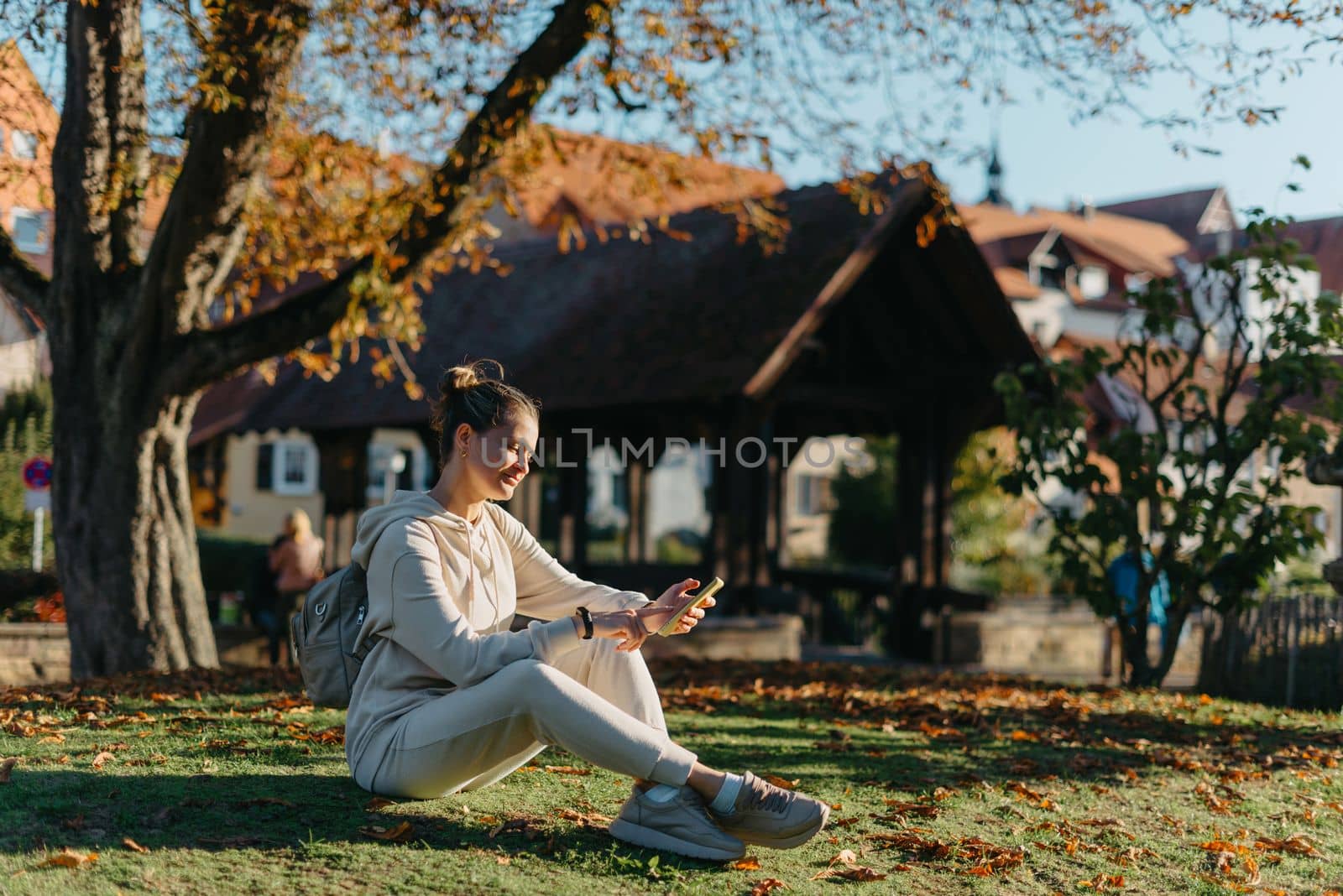 Young Fashionable Teenage Girl With Smartphone In Europian Park In Autumn Sitting At Smiling. Trendy Young Woman In Fall In Park Texting. Retouched, Vibrant Colors. Beautiful Blonde Teenage Girl Wearing Casual Modern Autumn Outfit Sitting In Park In Autumn. Retouched, Vibrant Colors, Brownish Tones. by Andrii_Ko