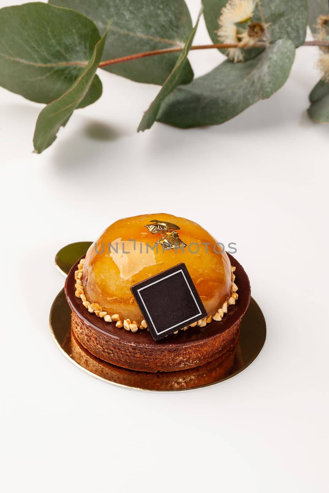 Crispy shortbread tartlet filled with almond cream topped with chocolate and caramelized apple decorated with nut crumbs on white surface with green flower branch in background