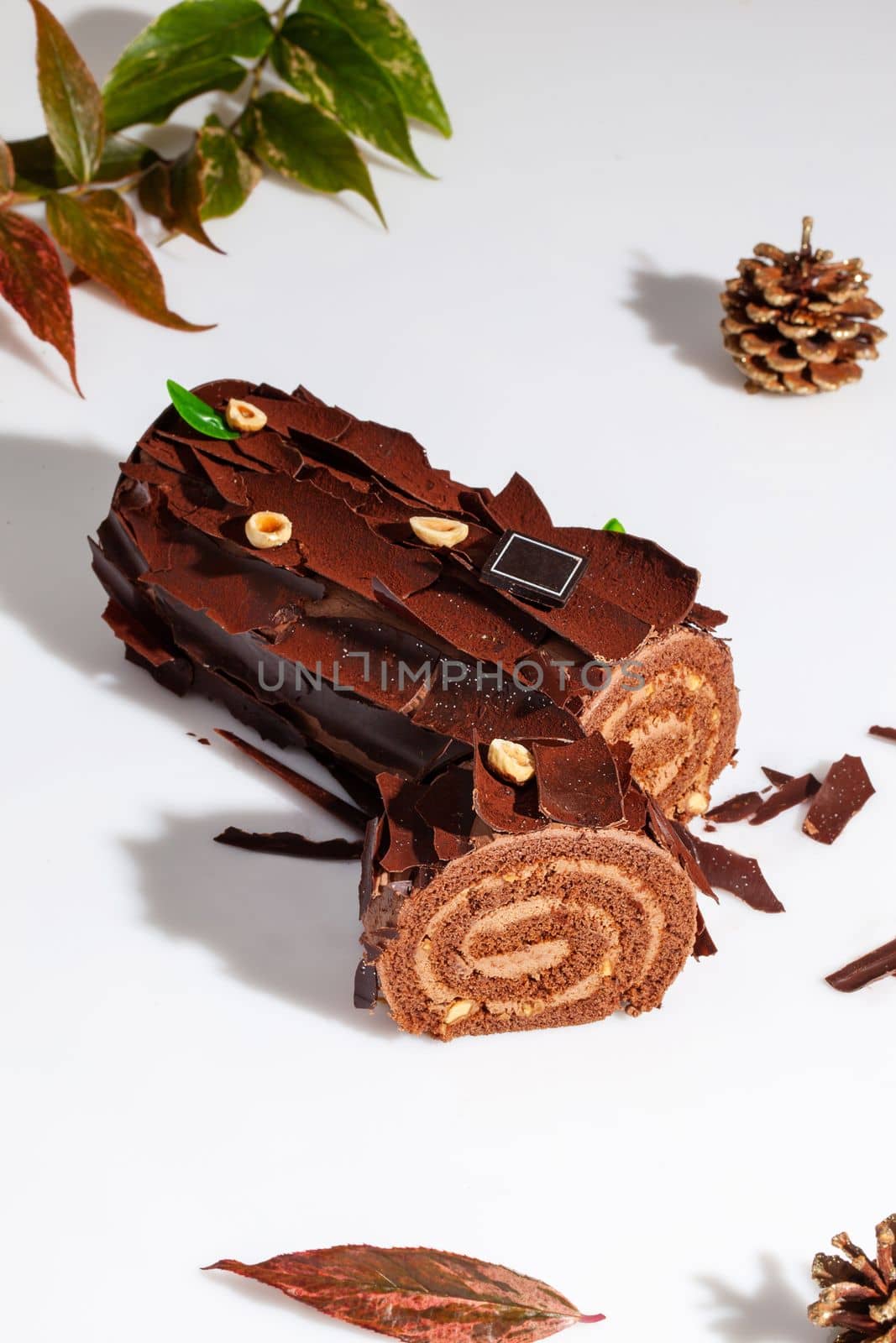 Sliced log-shaped sweet chocolate roll with tender buttercream filling, decorated with chocolate chips and hazelnuts on white background with gilded fir cone. Popular festive pastries concept