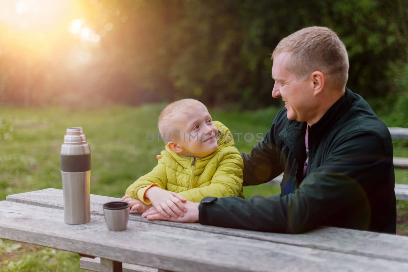 Happy Family: Father And Child Boy Son Playing And Laughing In Autumn Park, Sitting On Wooden Bench And Table. Father And Little Kid Having Fun Outdoors, Playing Together. Father And Son Sitting On A Bench And Talking. Thermos by Andrii_Ko