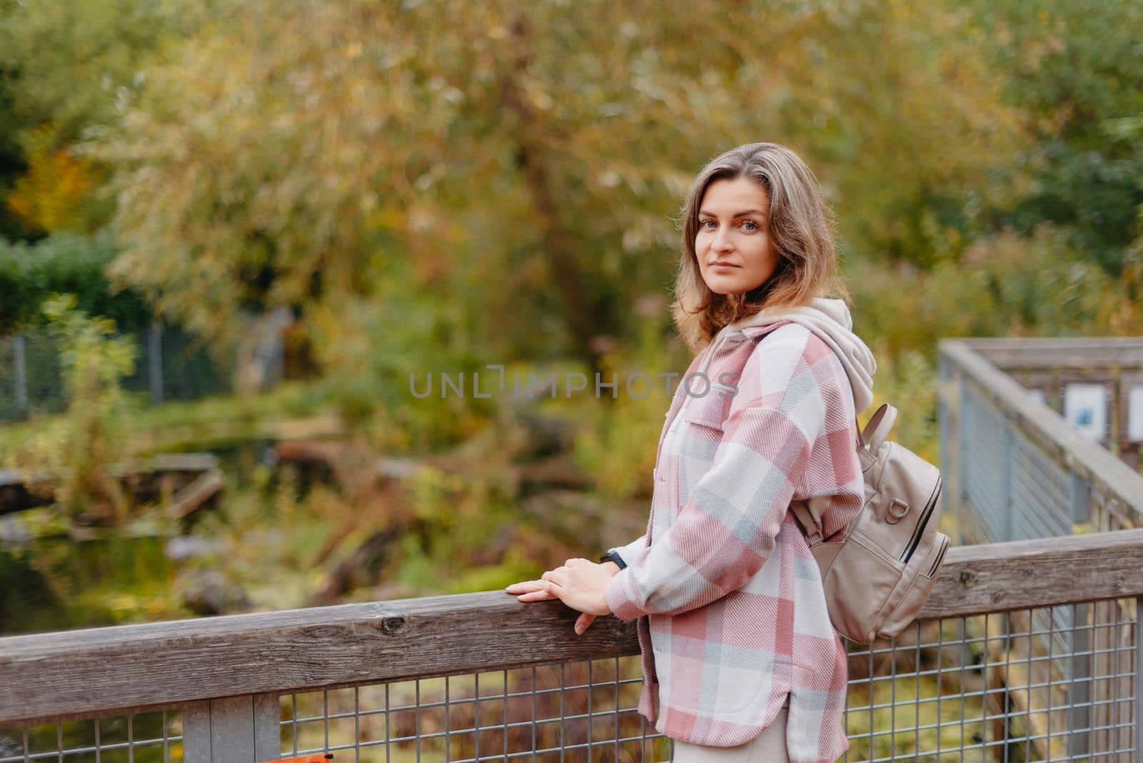 Portrait of cute young woman in casual wear in autumn, standing on bridge against background of an autumn Park and river. Pretty female walking in Park in golden fall. Copy space. smiling girl in the park standing on wooden bridge and looking at the camera in autumn season