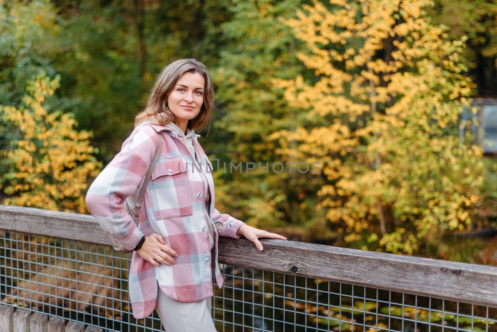 Portrait of cute young woman in casual wear in autumn, standing on bridge against background of an autumn Park and river. Pretty female walking in Park in golden fall. Copy space. smiling girl in the park standing on wooden bridge and looking at the camera in autumn season