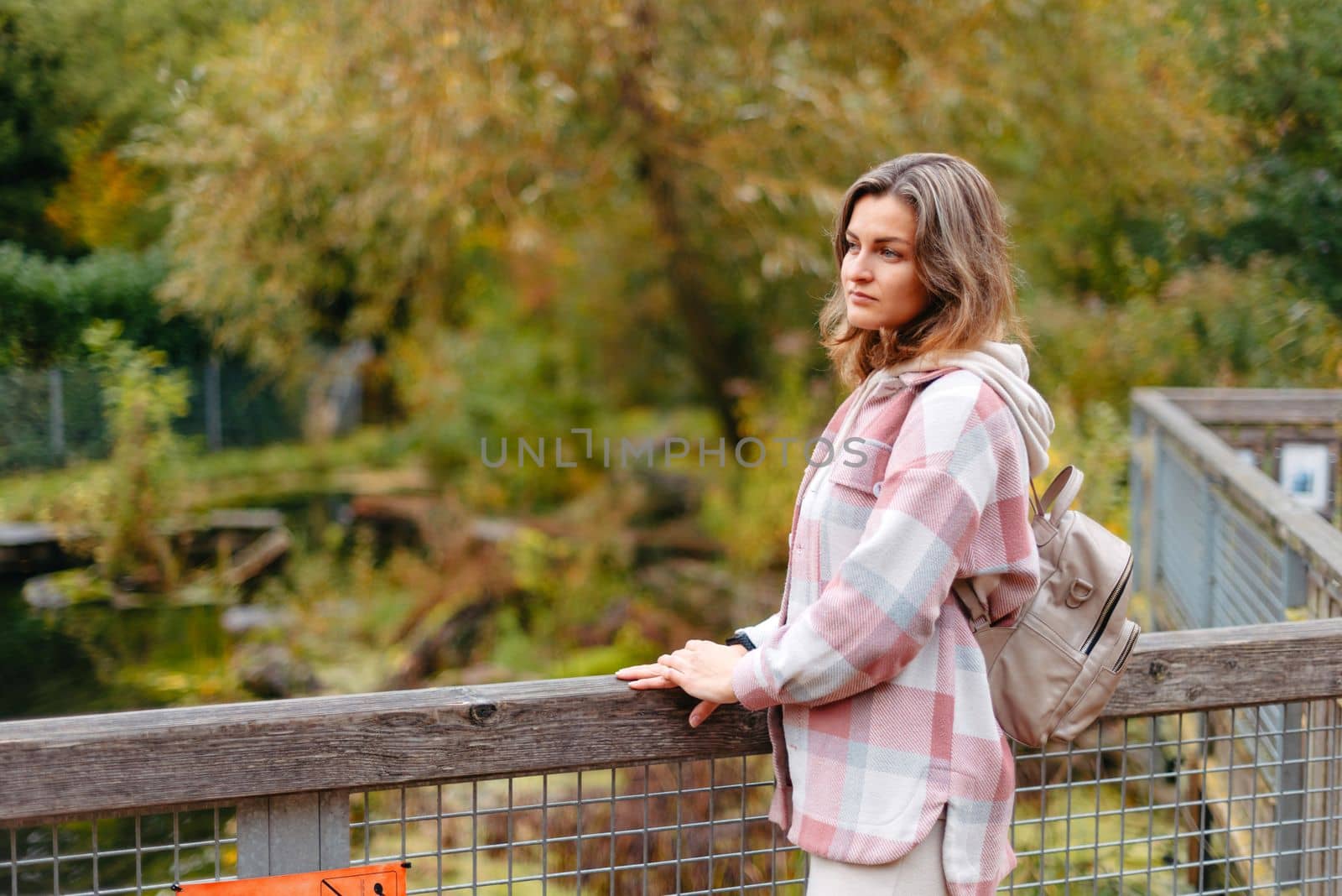 Portrait Of Cute Young Woman In Casual Wear In Autumn, Standing On Bridge Against Background Of An Autumn Park And River. Pretty Female Walking In Park In Golden Fall. Copy Space. Smiling Girl In The Park Standing On Wooden Bridge And Looking At The Camera In Autumn Season by Andrii_Ko