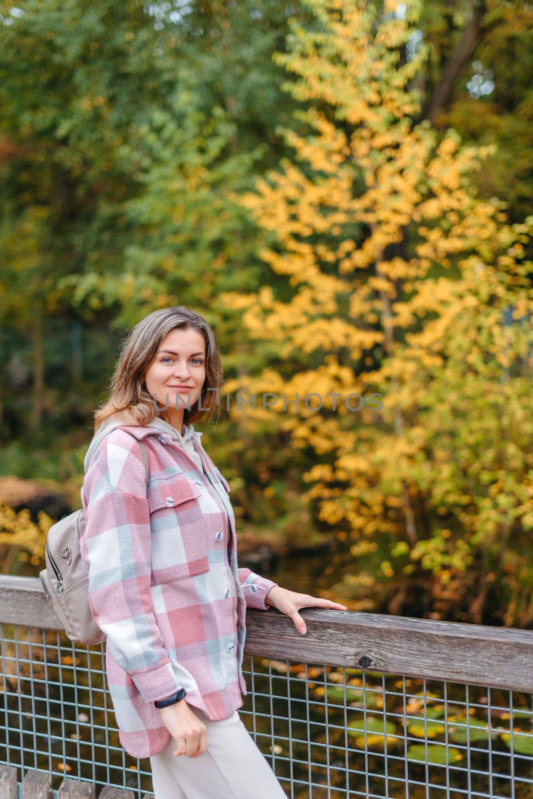 Portrait of cute young woman in casual wear in autumn, standing on bridge against background of an autumn Park and river. Pretty female walking in Park in golden fall. Copy space. smiling girl in the park standing on wooden bridge and looking at the camera in autumn season