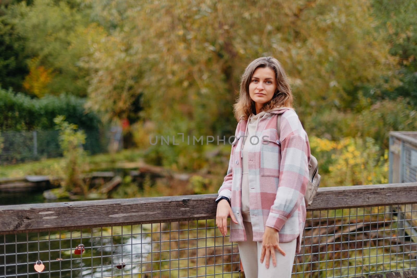 Portrait Of Cute Young Woman In Casual Wear In Autumn, Standing On Bridge Against Background Of An Autumn Park And River. Pretty Female Walking In Park In Golden Fall. Copy Space. Smiling Girl In The Park Standing On Wooden Bridge And Looking At The Camera In Autumn Season by Andrii_Ko