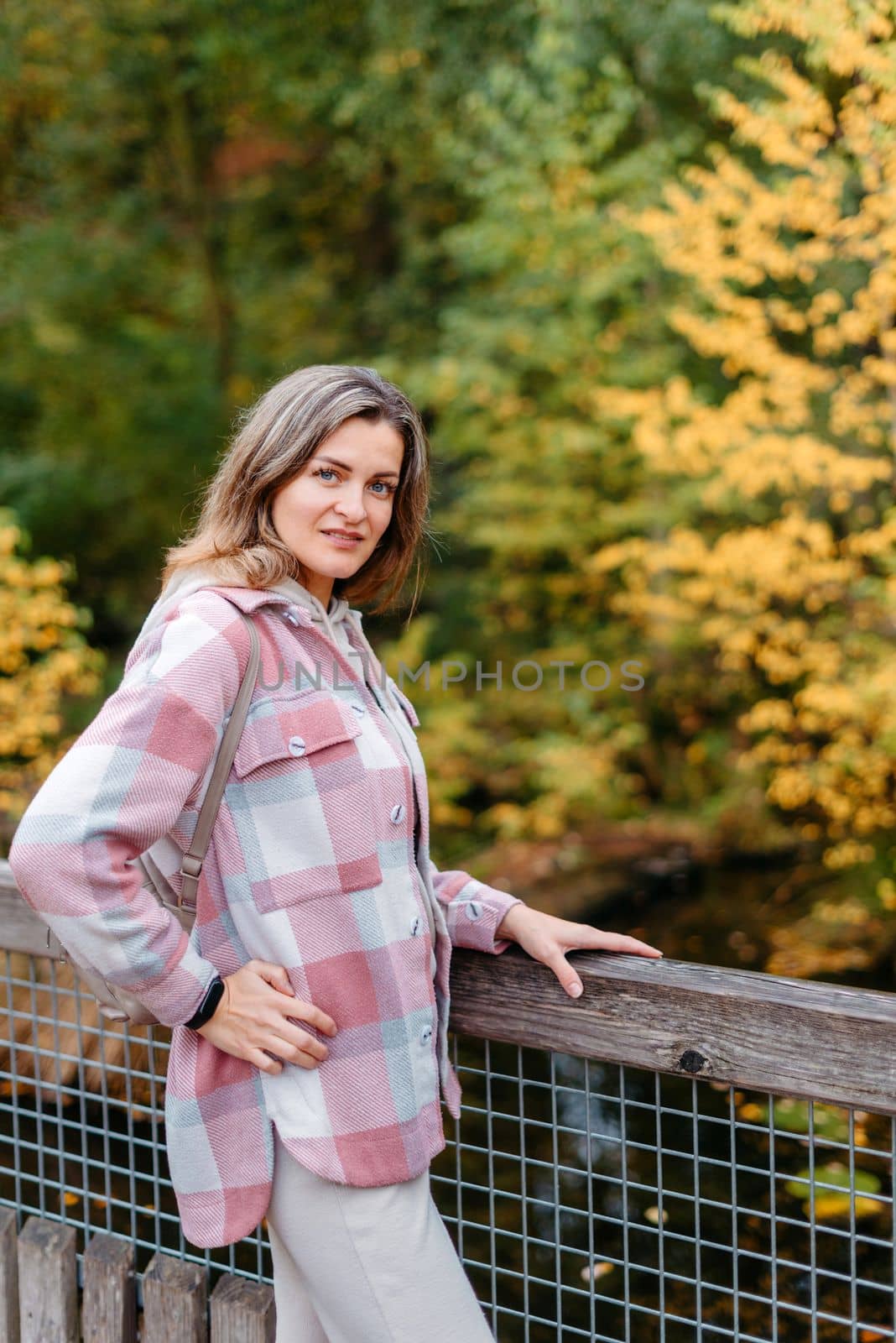 Portrait Of Cute Young Woman In Casual Wear In Autumn, Standing On Bridge Against Background Of An Autumn Park And River. Pretty Female Walking In Park In Golden Fall. Copy Space. Smiling Girl In The Park Standing On Wooden Bridge And Looking At The Camera In Autumn Season by Andrii_Ko