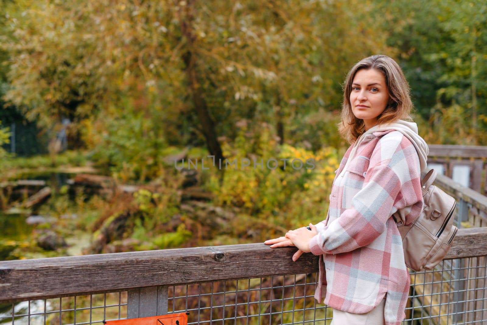 Portrait of cute young woman in casual wear in autumn, standing on bridge against background of an autumn Park and river. Pretty female walking in Park in golden fall. Copy space. smiling girl in the park standing on wooden bridge and looking at the camera in autumn season