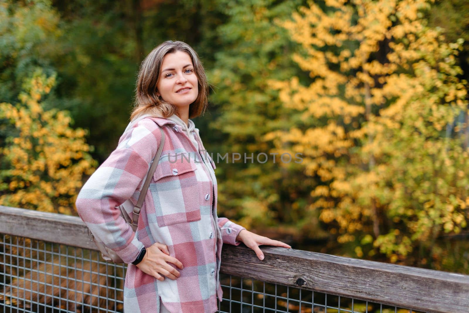 Portrait of cute young woman in casual wear in autumn, standing on bridge against background of an autumn Park and river. Pretty female walking in Park in golden fall. Copy space. smiling girl in the park standing on wooden bridge and looking at the camera in autumn season